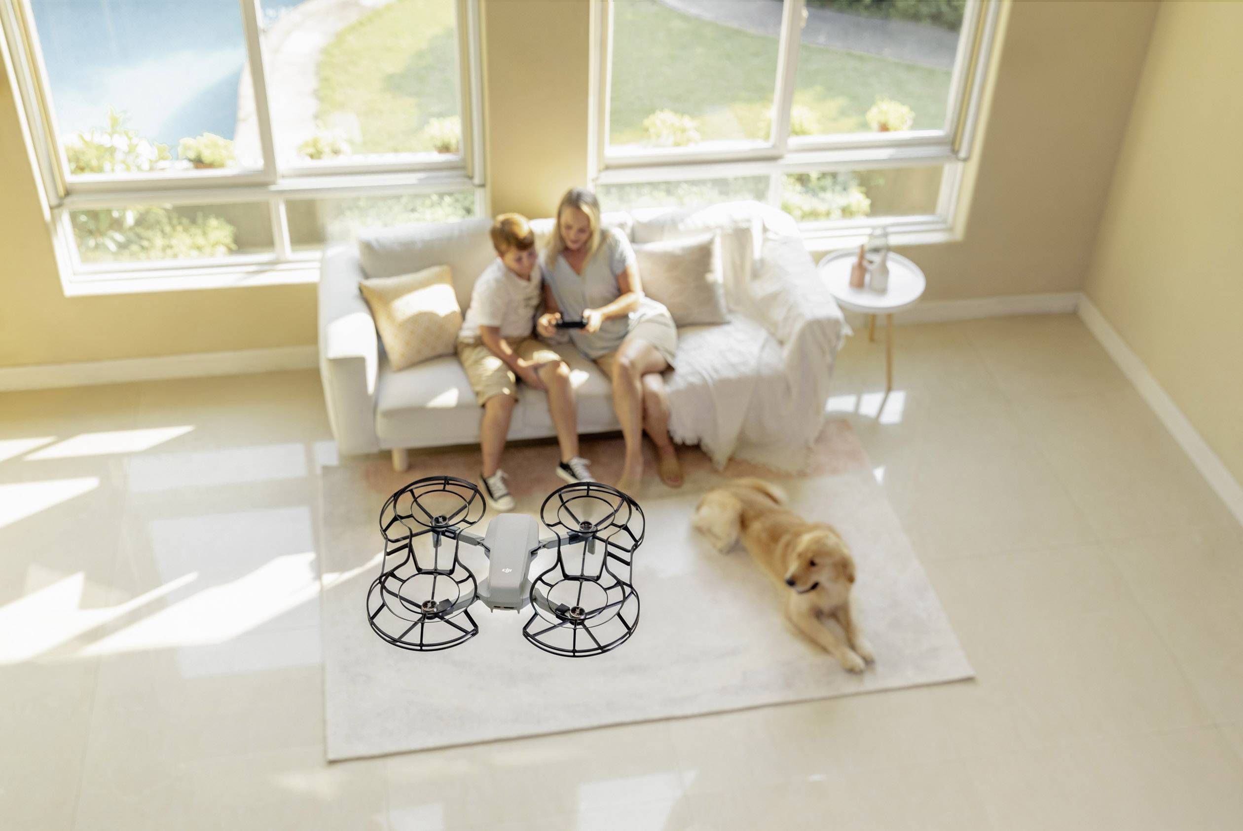 Une femme et un enfant sont assis sur un canapé et jouent avec un drone. Un chien est allongé sur le sol. Pièce bien éclairée avec vue sur l'extérieur.