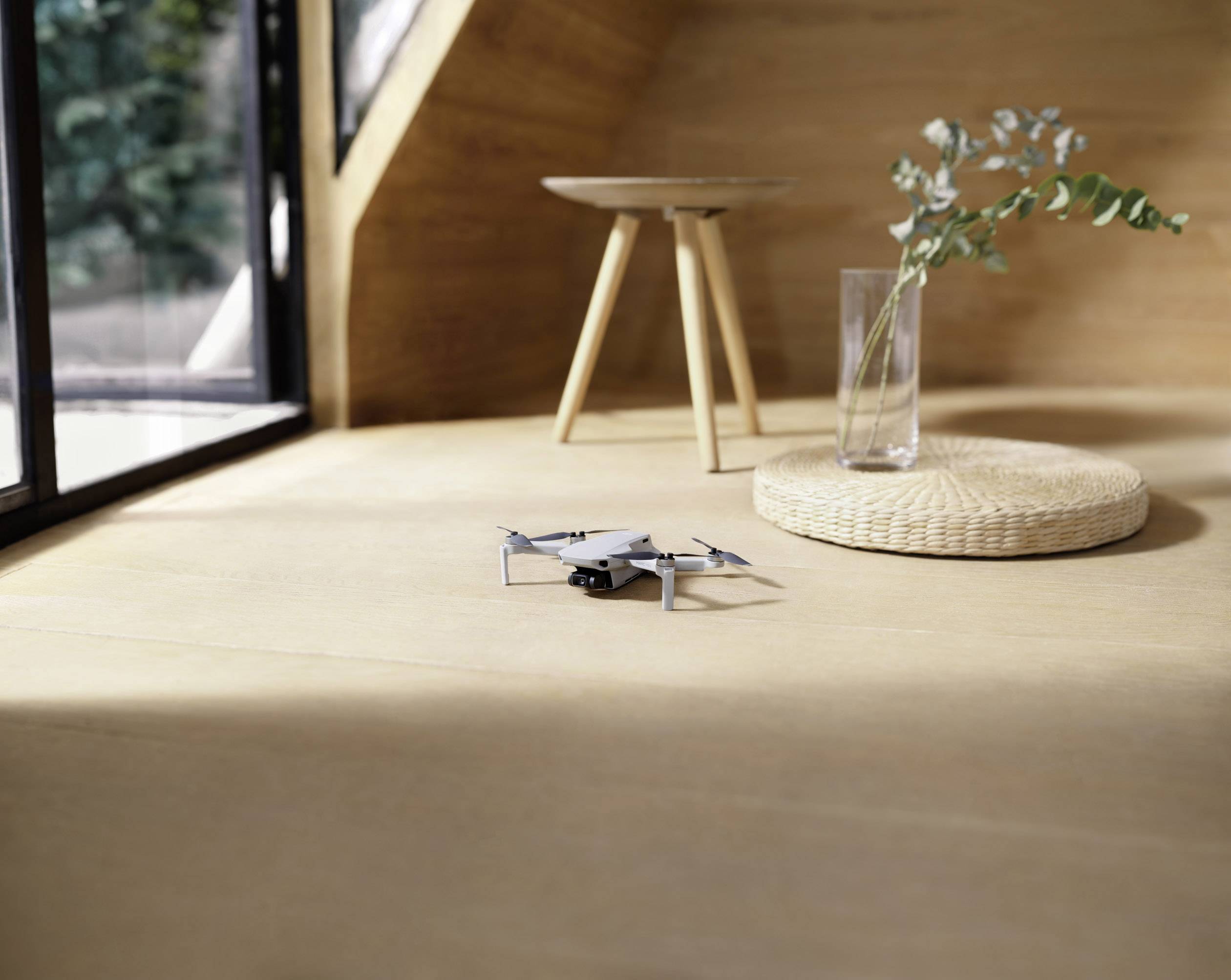 Un petit drone est posé sur une table dans une pièce ensoleillée, à côté se trouve une chaise et un vase en verre avec des plantes.