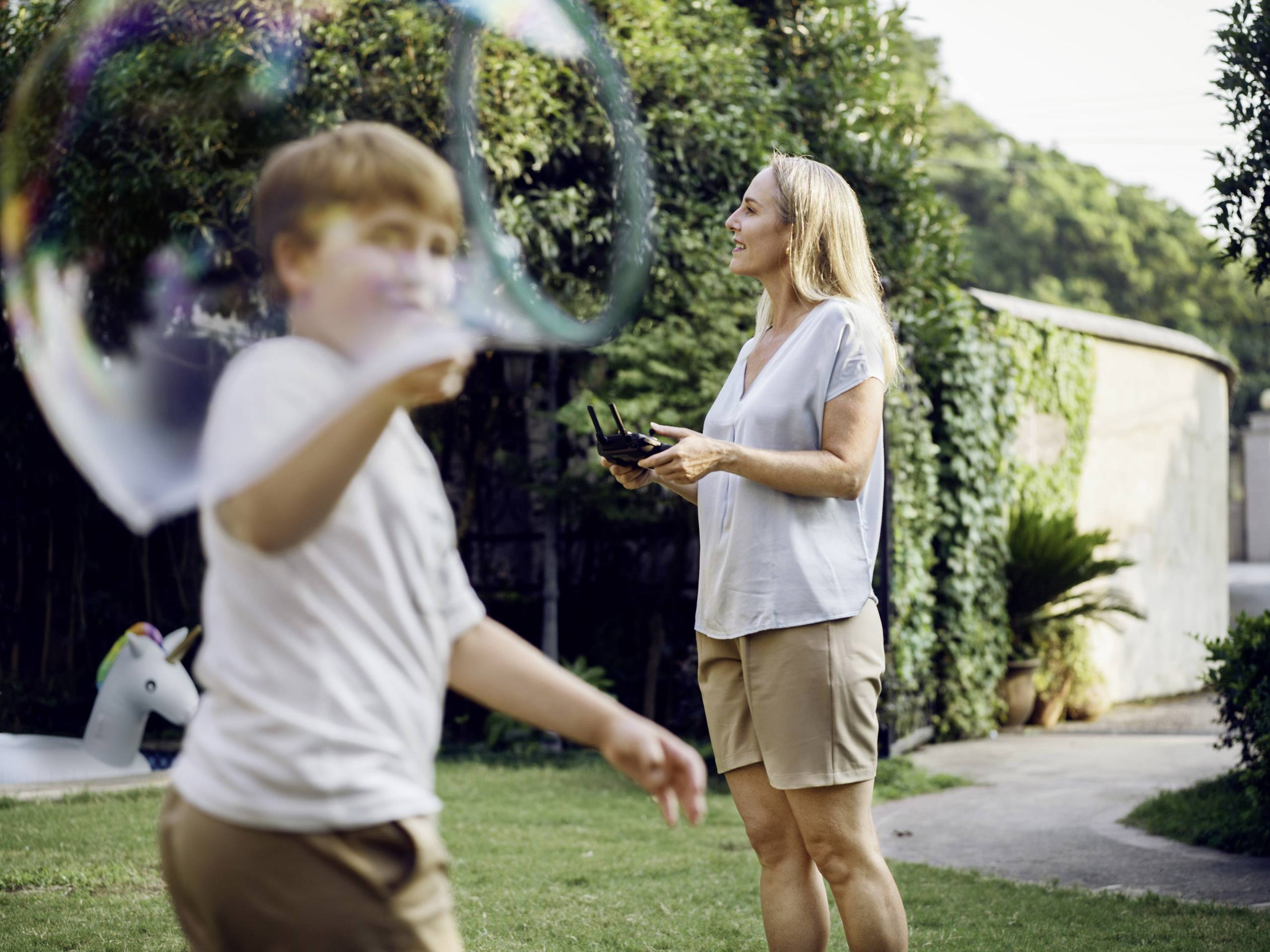 Une femme dans un jardin fait voler un drone, tandis qu'un enfant fait des bulles de savon. À l'arrière-plan, on aperçoit une licorne gonflable.