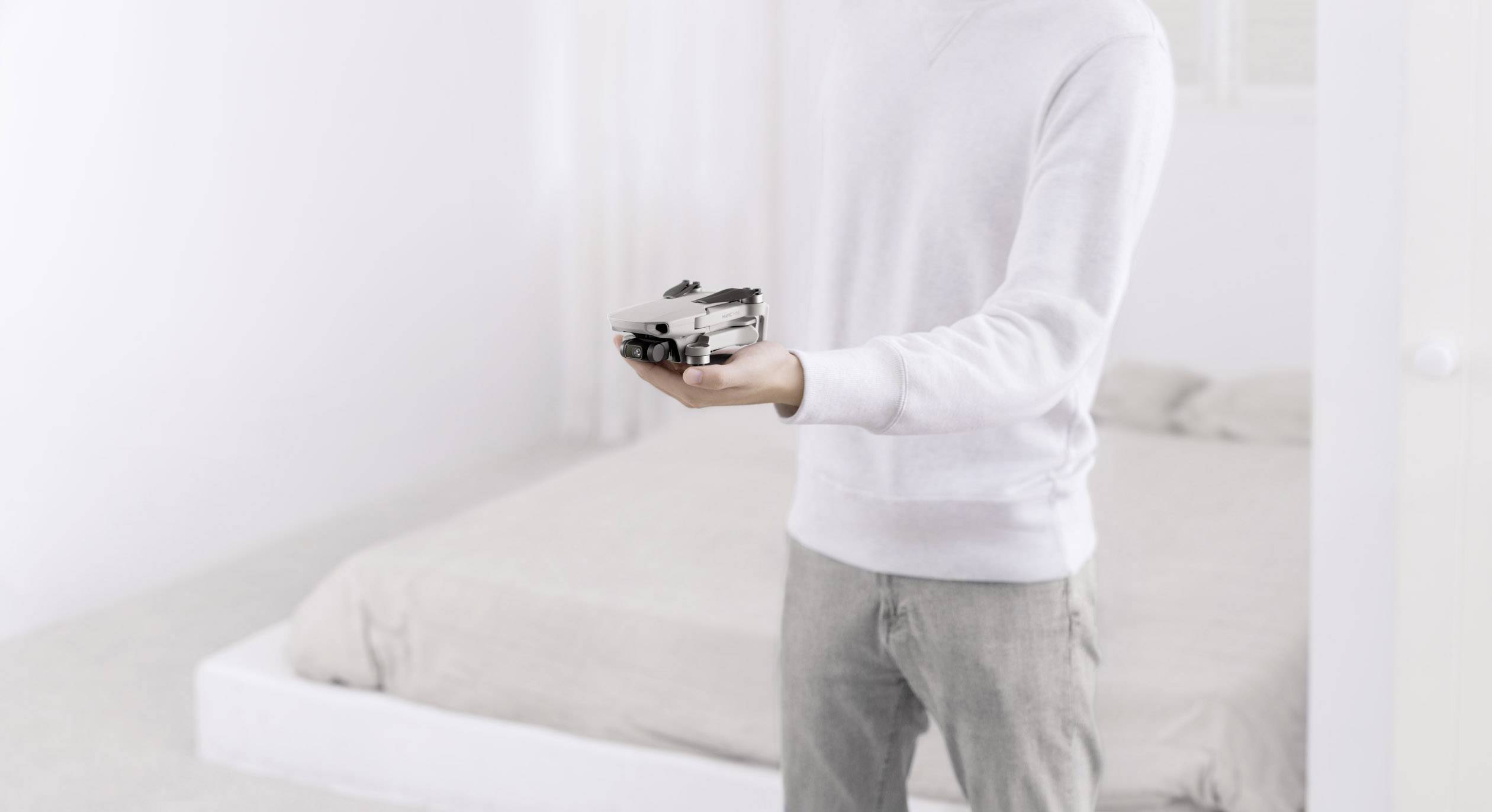 Une personne portant un pull blanc tient un drone gris dans sa main, debout dans une chambre lumineuse devant un lit.