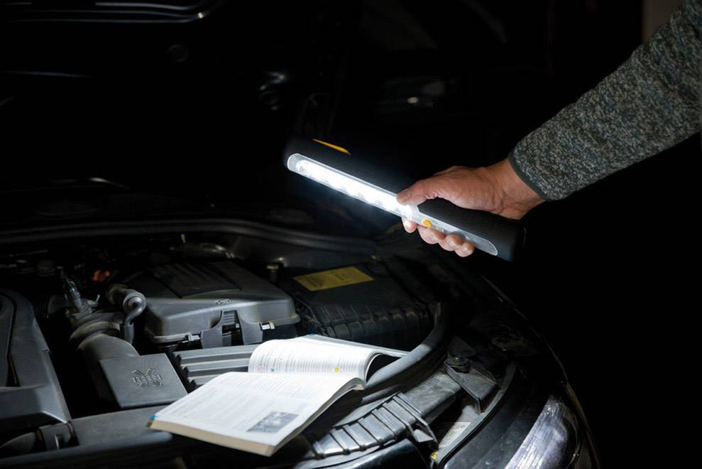 Une personne tient une lampe de poche au-dessus du capot ouvert d'une voiture. Un manuel est posé sur le moteur, la lumière illuminant la zone.