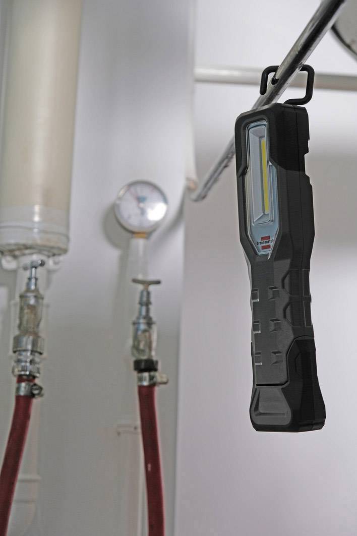Lumière LED portable suspendue à un tube dans une salle technique, entourée de tuyaux et d'un manomètre, illustrant un environnement industriel.