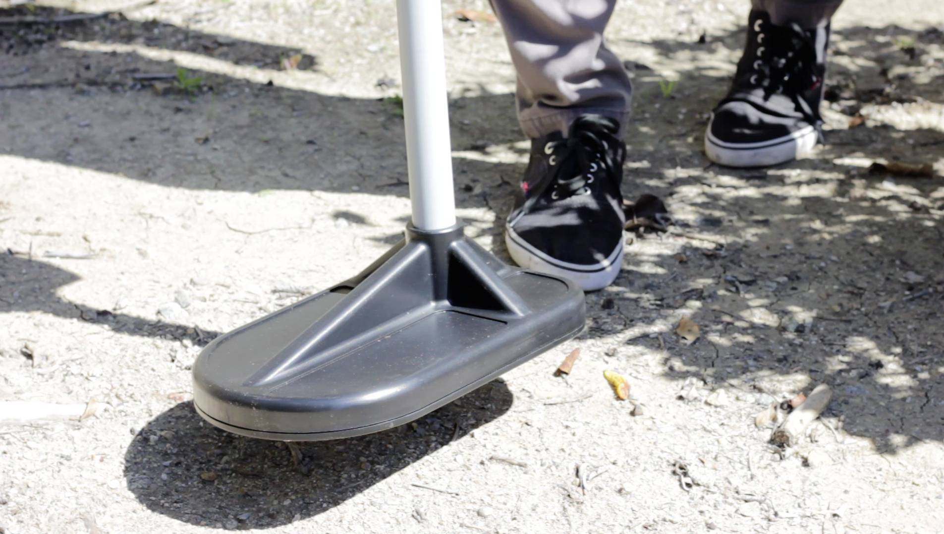 Une personne nettoie le sol avec un balai. On voit ses chaussures noires sur une surface en béton ensoleillée.