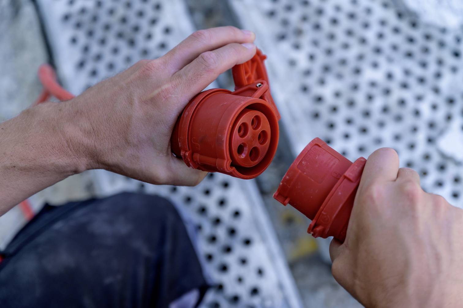 Deux mains tiennent une fiche électrique rouge et sa prise correspondante, prêtes à être connectées. Arrière-plan : grille métallique.