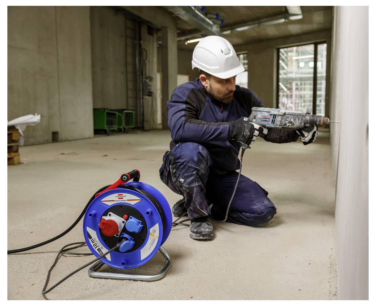 Ouvrier du bâtiment en casque de protection s'agenouille en perçant un mur en béton, avec une bobine de câble bleu au premier plan.