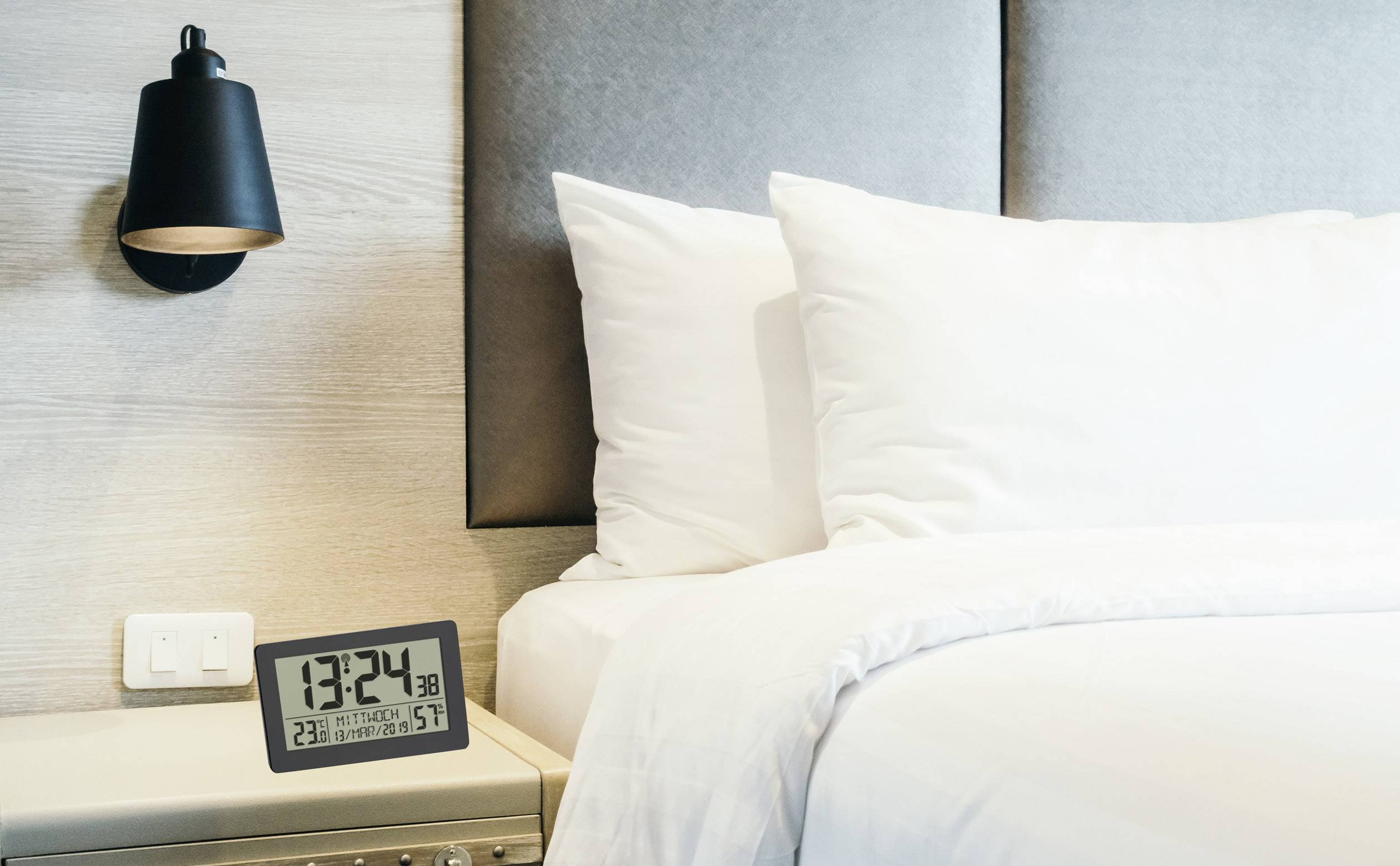 Horloge numérique affichant 13:24 sur la table de nuit à côté du lit avec des oreillers blancs et une couverture ; lampe moderne fixée au mur.