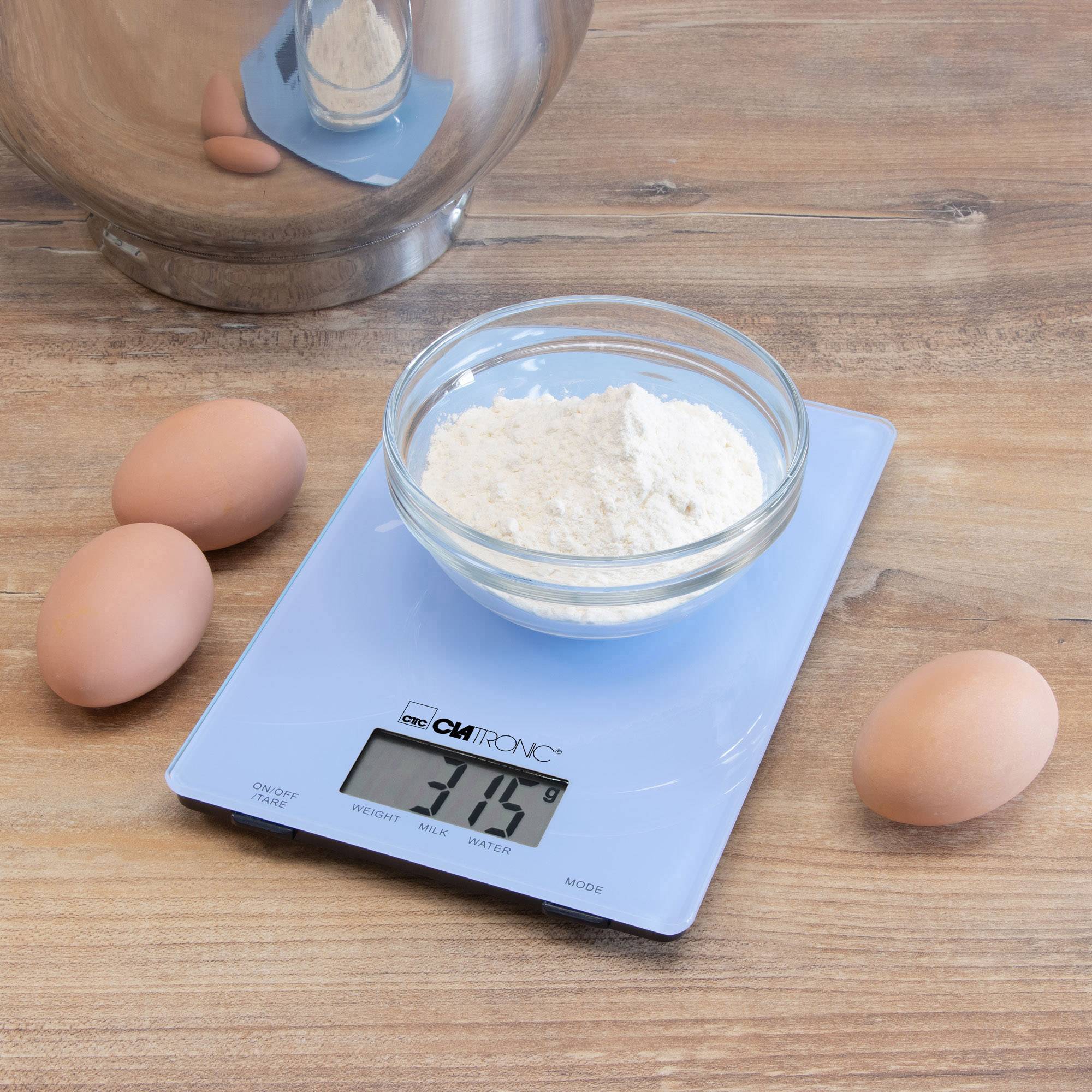 Une balance de cuisine numérique affiche 315 g de farine dans un bol en verre. À côté, quatre œufs bruns sont posés sur une surface en bois.