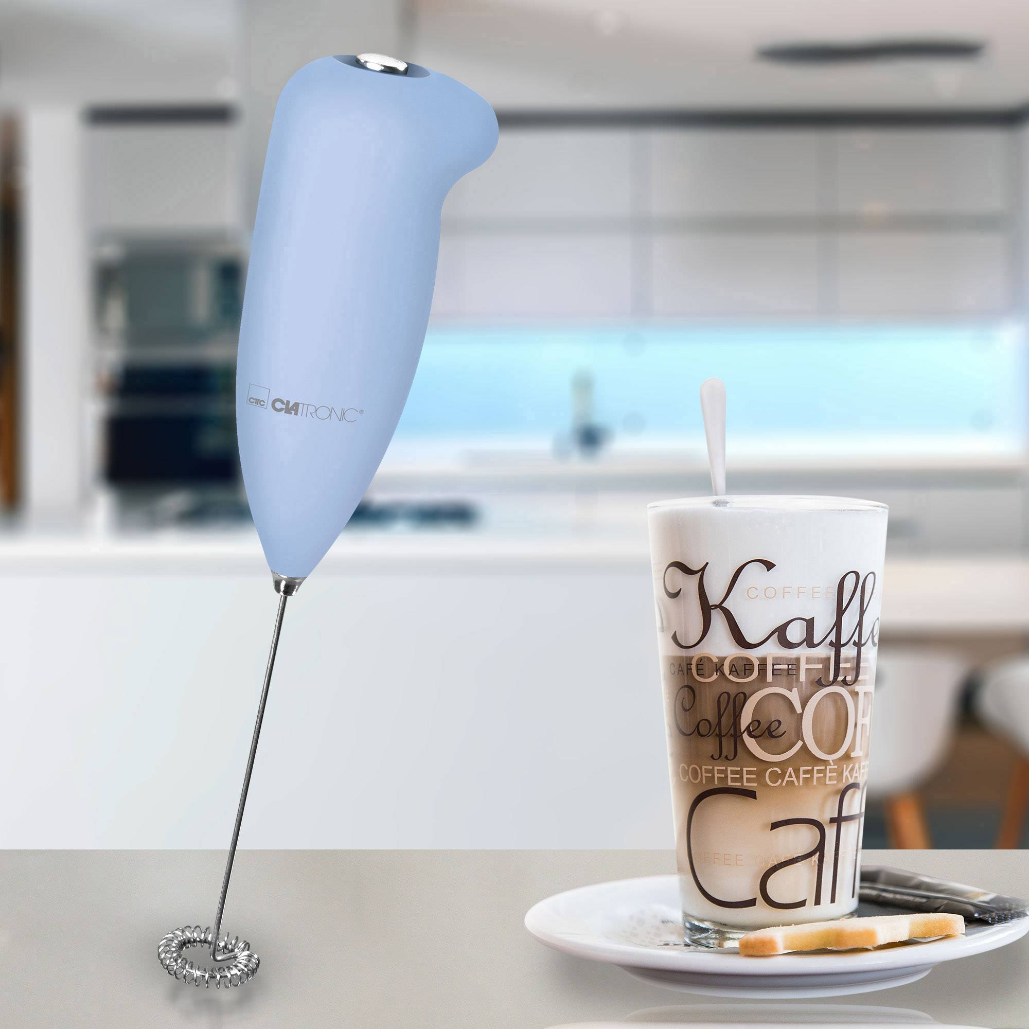 Un mousseur à lait bleu est placé dans une cuisine moderne à côté d'un verre de Latte Macchiato, sur lequel est écrit plusieurs fois le mot 'coffee'.