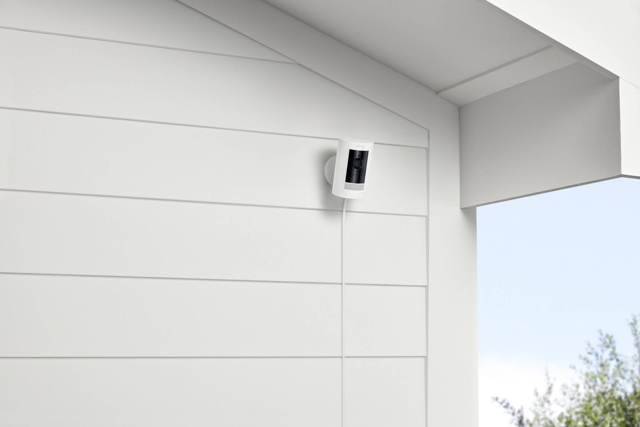 Caméra de sécurité blanche fixée sur le mur extérieur d'une maison, installée sous l'avant-toit. Des arbres sont visibles en arrière-plan.