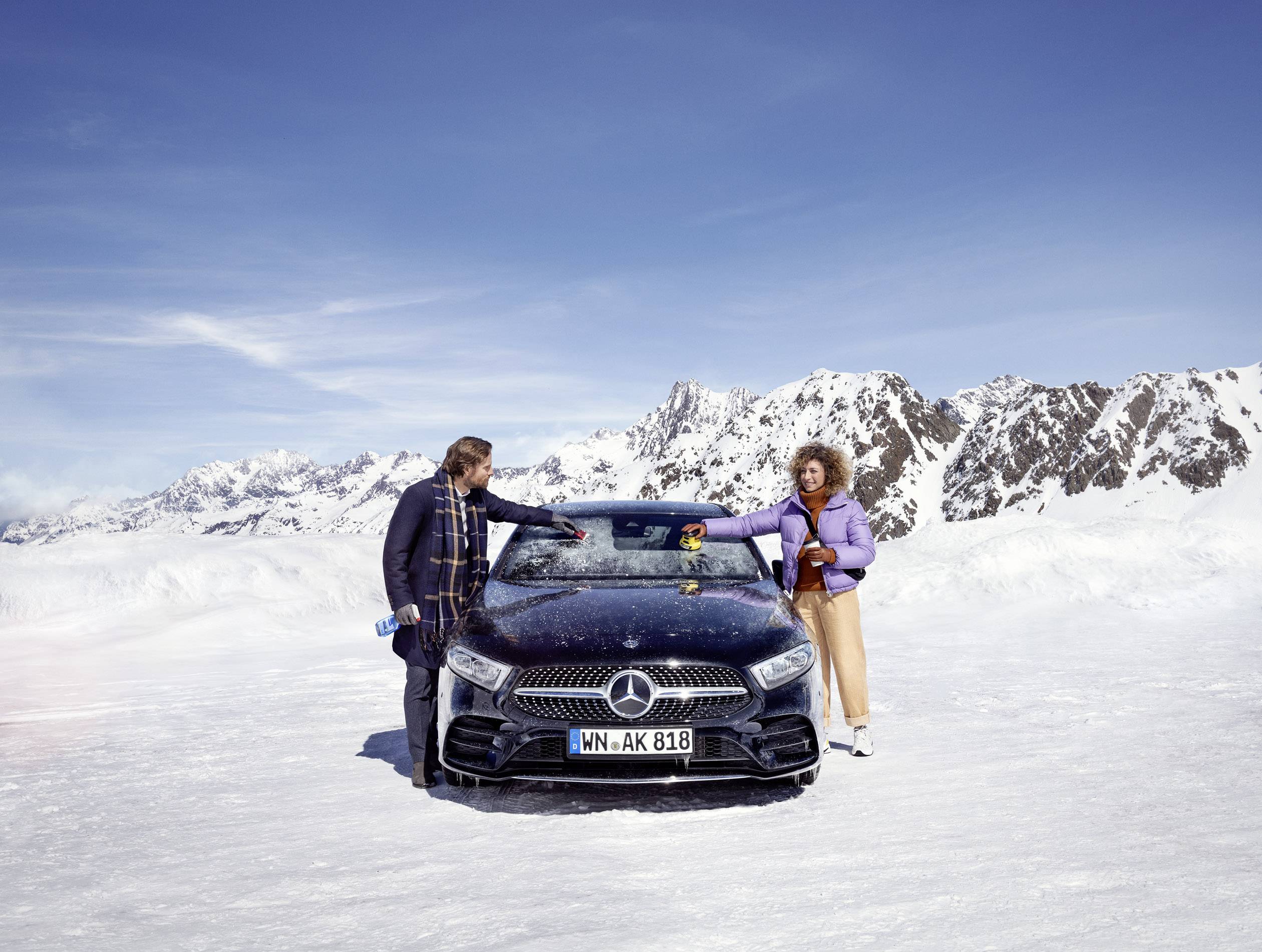 Deux personnes se tiennent devant une Mercedes dans un paysage montagneux enneigé. Une personne porte une veste violette, l'autre est vêtue de vêtements rayés.