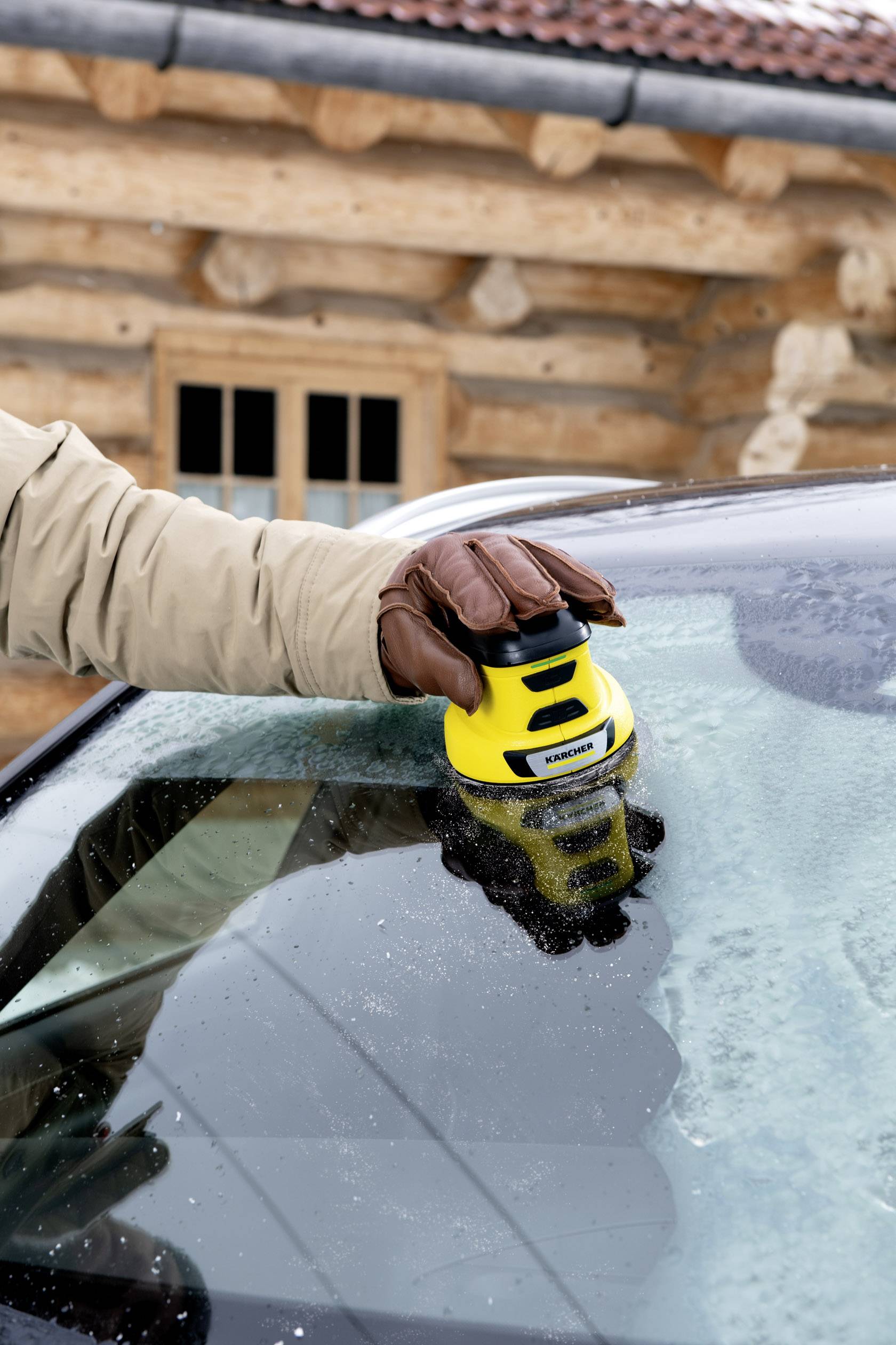 Une personne retire de la glace du pare-brise d'une voiture à l'aide d'un grattoir à glace jaune devant une maison en bois en hiver.