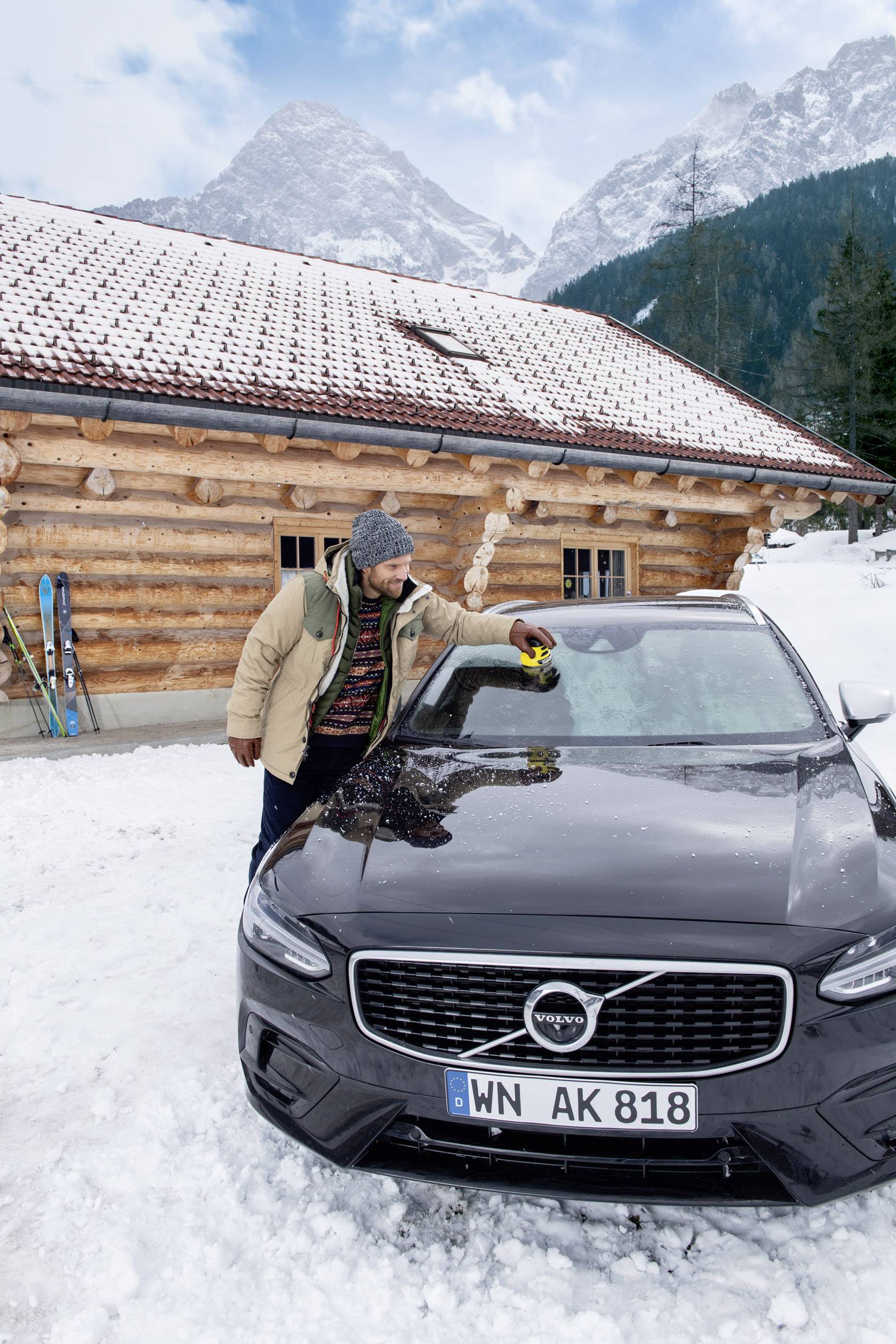 Un homme vêtu de vêtements d'hiver essuie la neige d'une voiture noire devant un chalet en bois et un paysage montagneux enneigé.