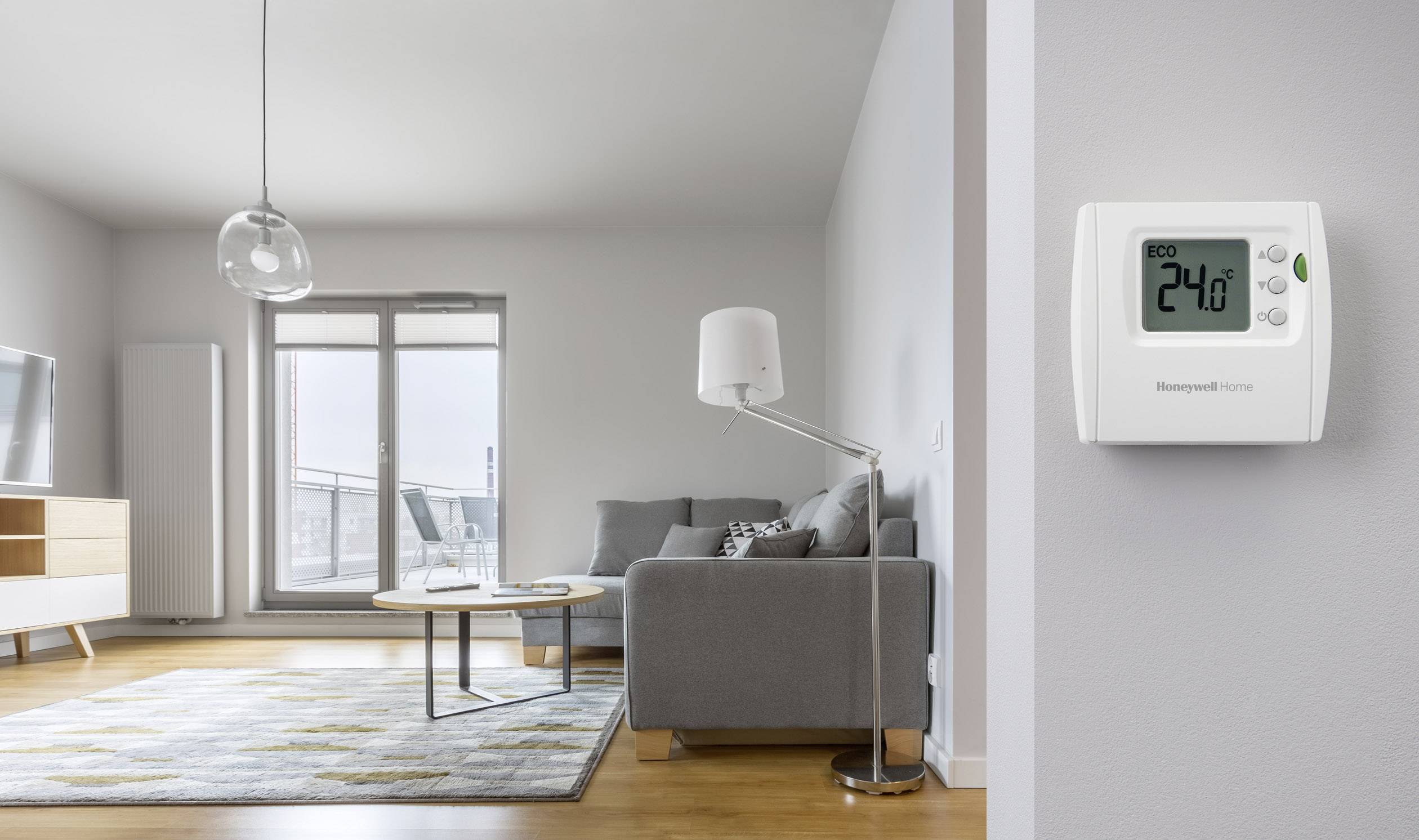 Salon avec un canapé gris, une lampe et un tapis. Un thermostat numérique est fixé au mur, affichant '24°C', en mode Eco. Balcon visible en arrière-plan.