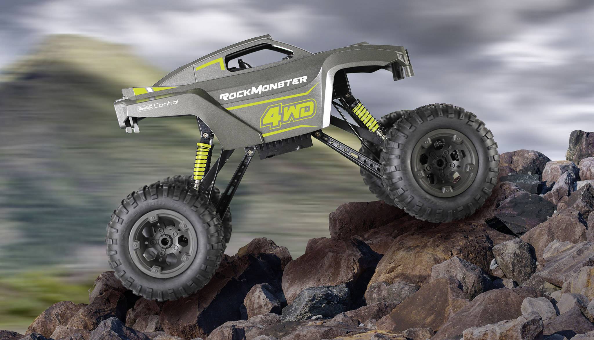 Un véhicule télécommandé tout-terrain avec de grandes roues et suspension circule sur un terrain rocailleux. Des montagnes floues se profilent en arrière-plan.