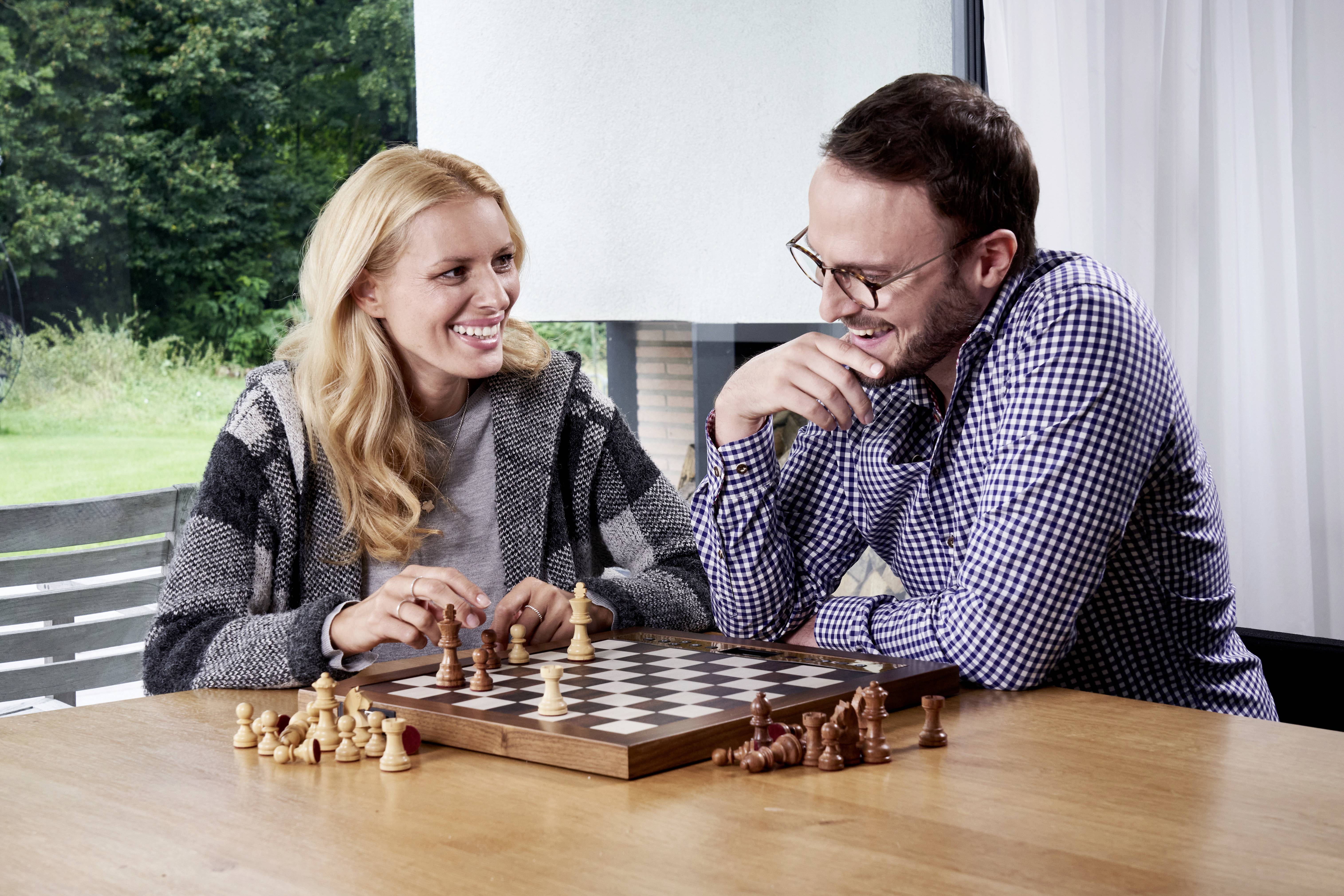 Deux personnes sont assises à une table, souriant et jouant aux échecs. Elles semblent détendues et concentrées sur la partie.