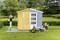 Une femme peint un petit cabanon bicolore en jaune, tandis qu'une autre se détend dans un transat. De l'herbe et des arbres sont visibles en arrière-plan.