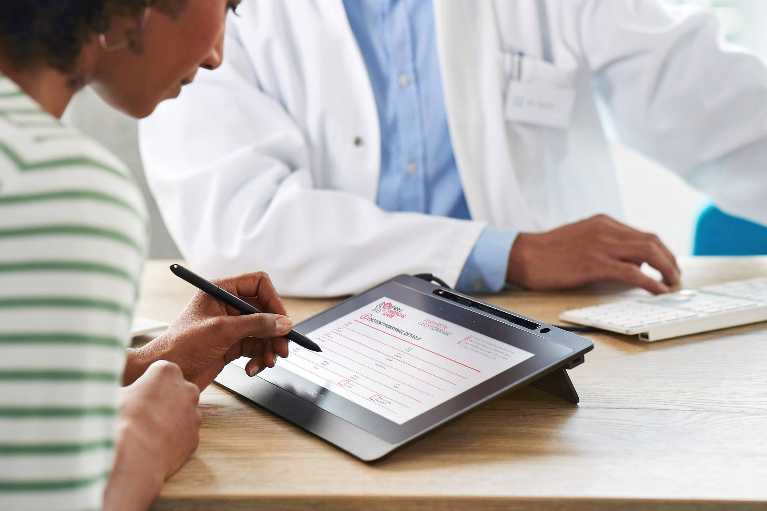 Une femme remplit un formulaire sur une tablette graphique pendant qu'un médecin est assis devant un ordinateur.