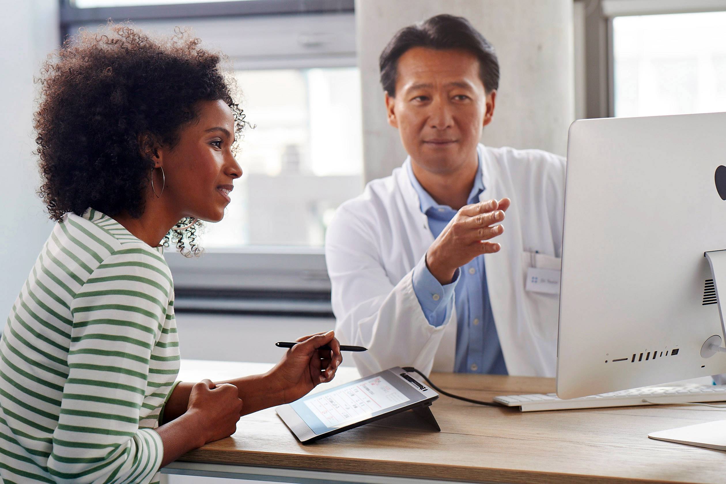 Un médecin explique quelque chose à une femme sur l'écran de l'ordinateur. Elle est assise en face de lui dans un bureau. Une tablette est posée sur la table.