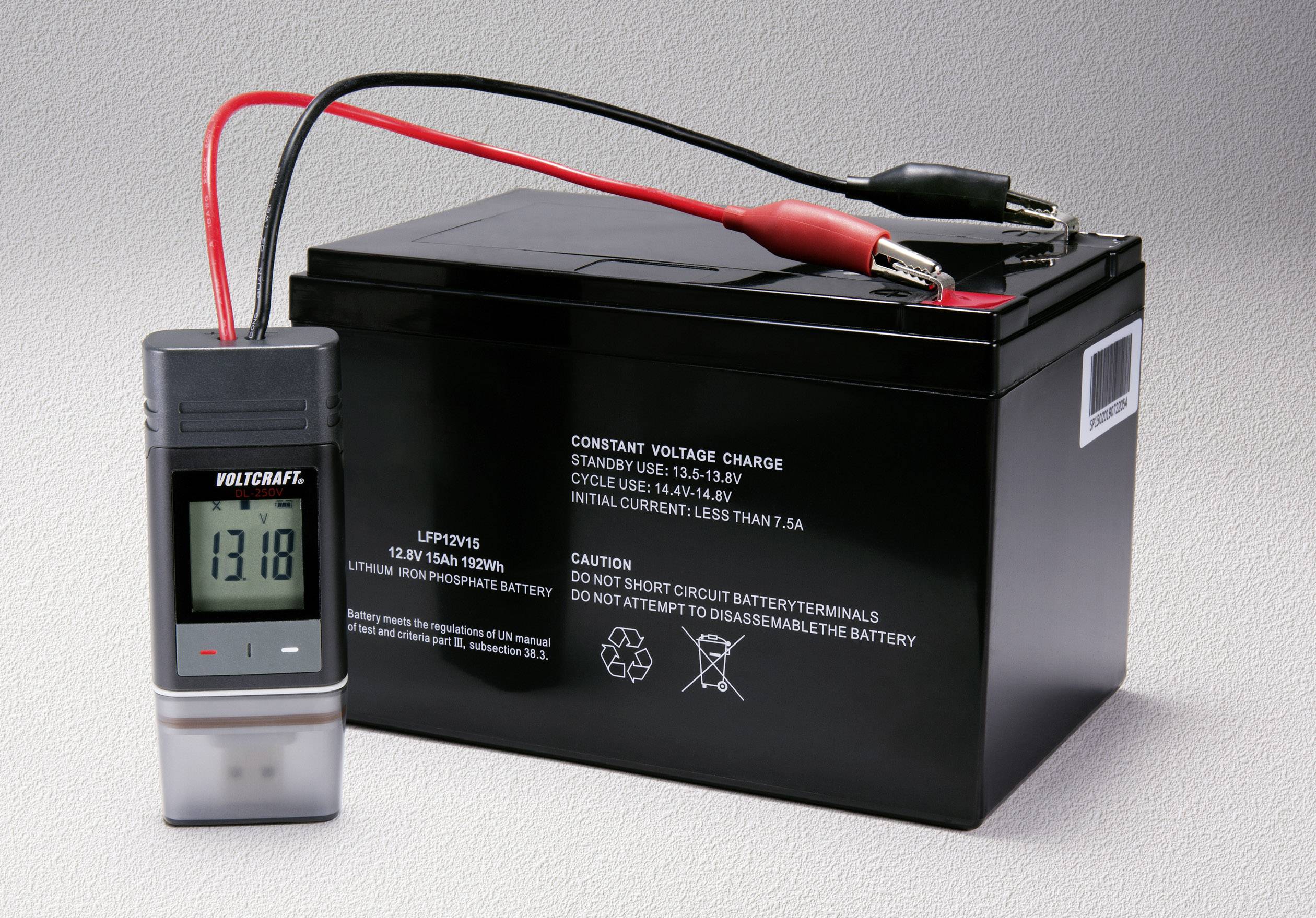 Un multimètre numérique affiche '13,18' et est connecté à une batterie de voiture noire. Des câbles rouge et noir mènent à l'appareil.