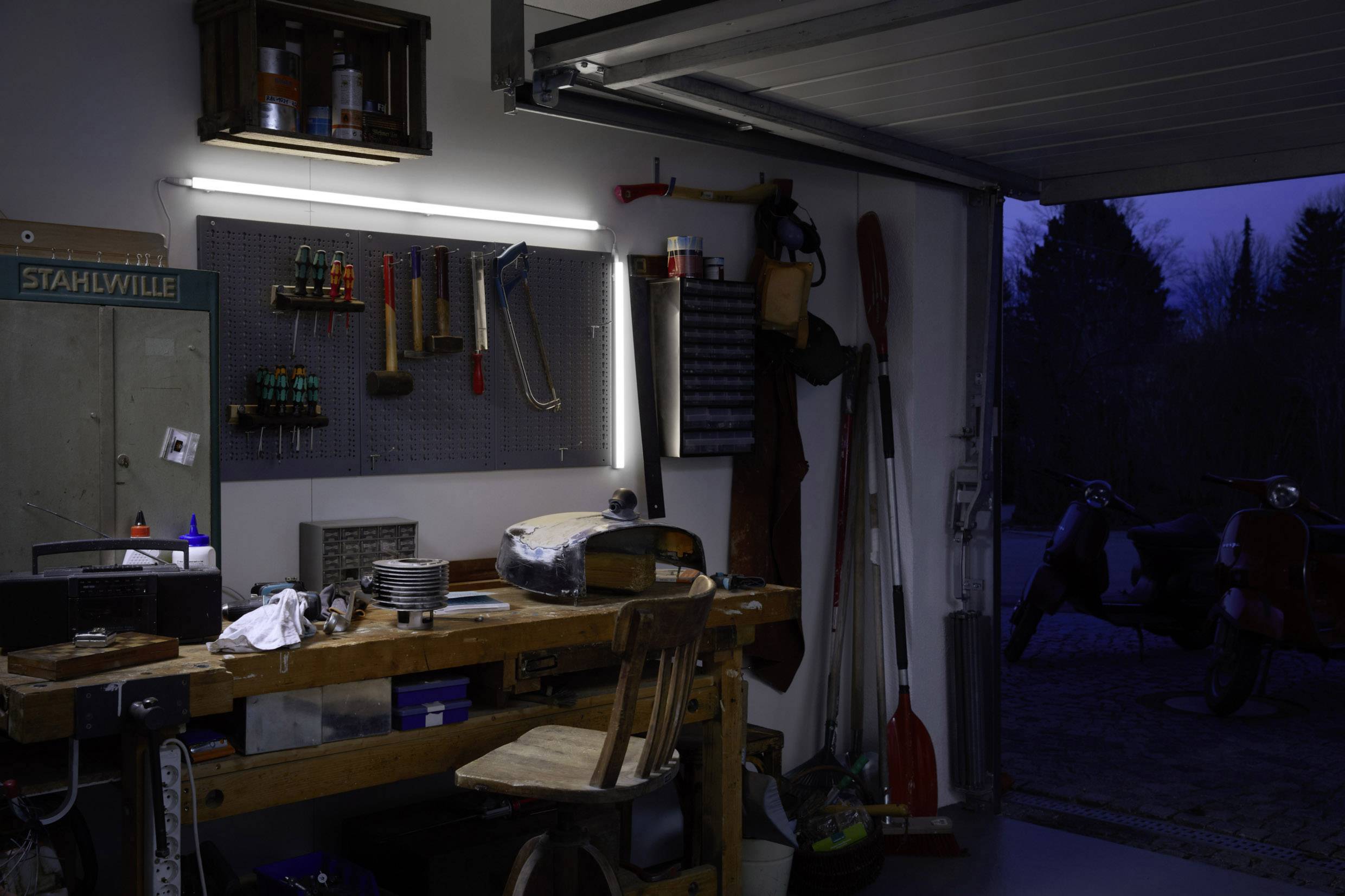 Un atelier avec des outils accrochés au mur, une table de travail en bois et deux scooters en arrière-plan au crépuscule.