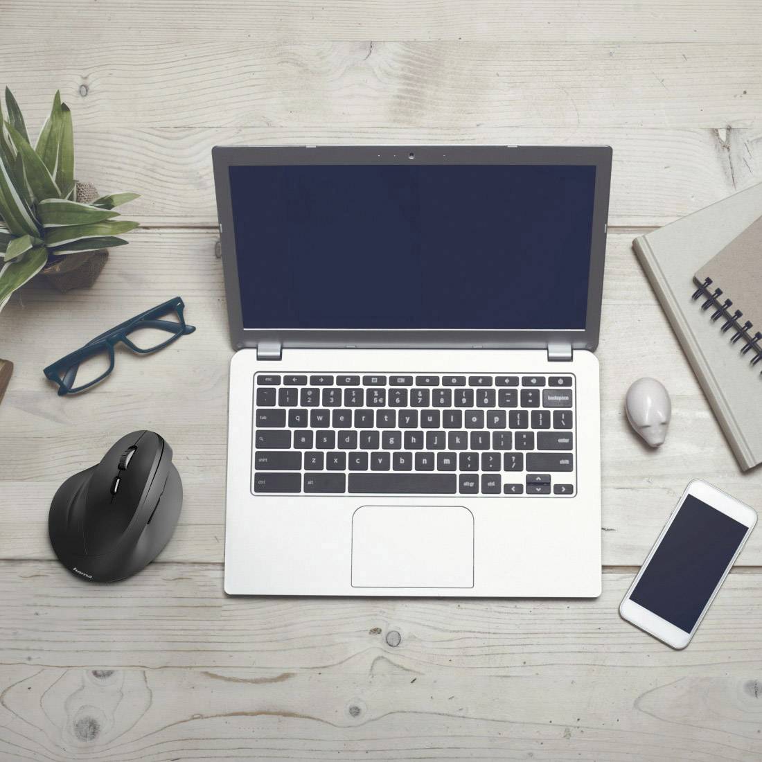 Un ordinateur portable sur une table en bois avec une souris, un téléphone portable, des lunettes, un carnet de notes et une plante.