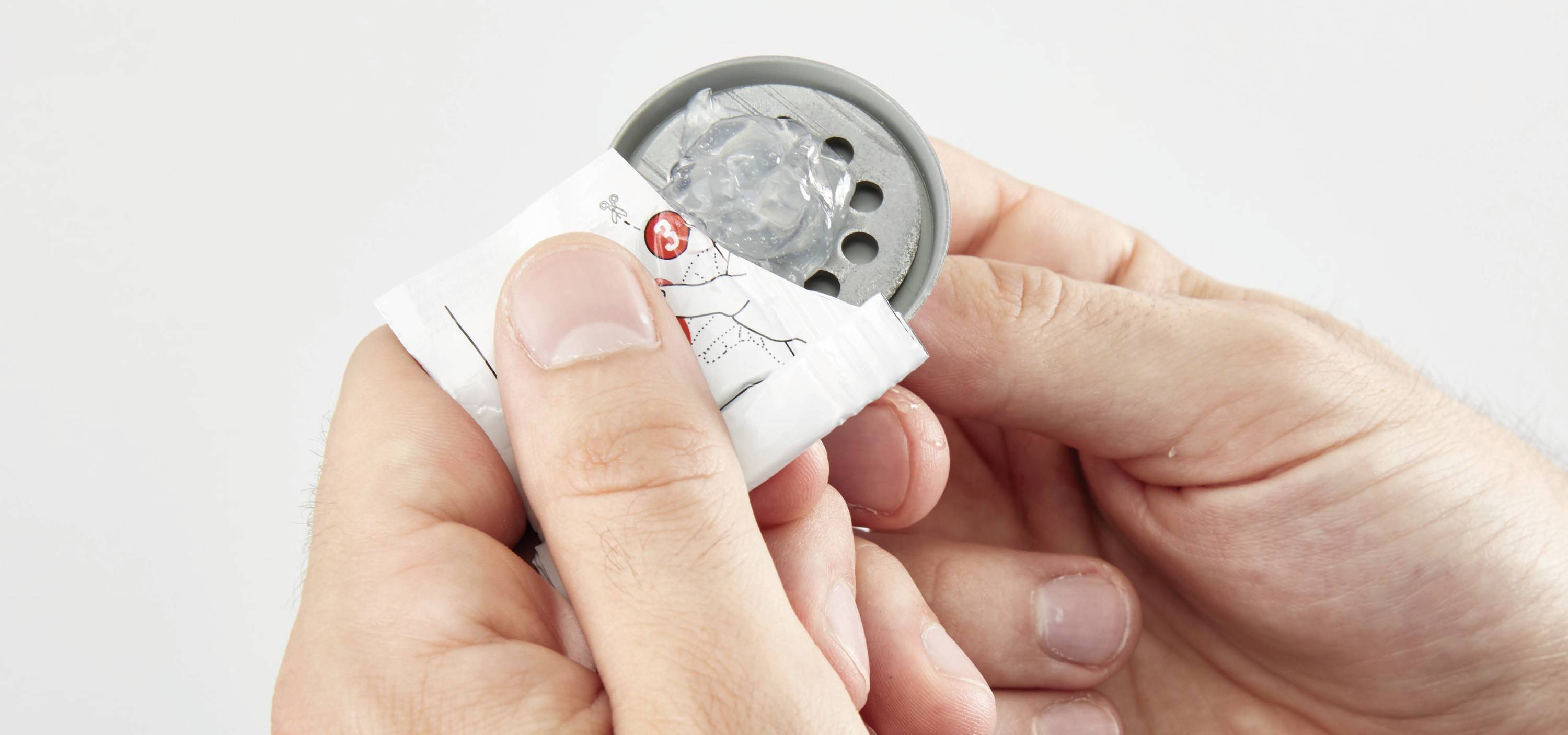 Deux mains ouvrent un emballage blanc pour libérer un objet gris ressemblant à du plastique.