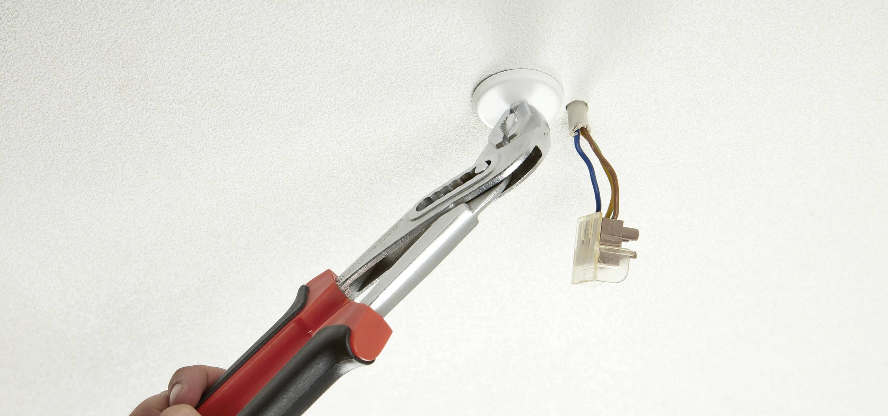 Une main tient une pince qui travaille sur un câble électrique suspendu à une ouverture au plafond.