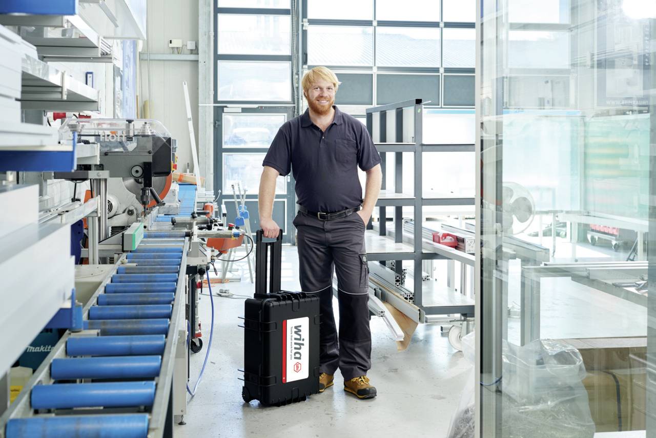 Une personne se tient dans un atelier avec des machines et une valise à roulettes portant l'inscription « wih? ». Un décor industriel se profile en arrière-plan.