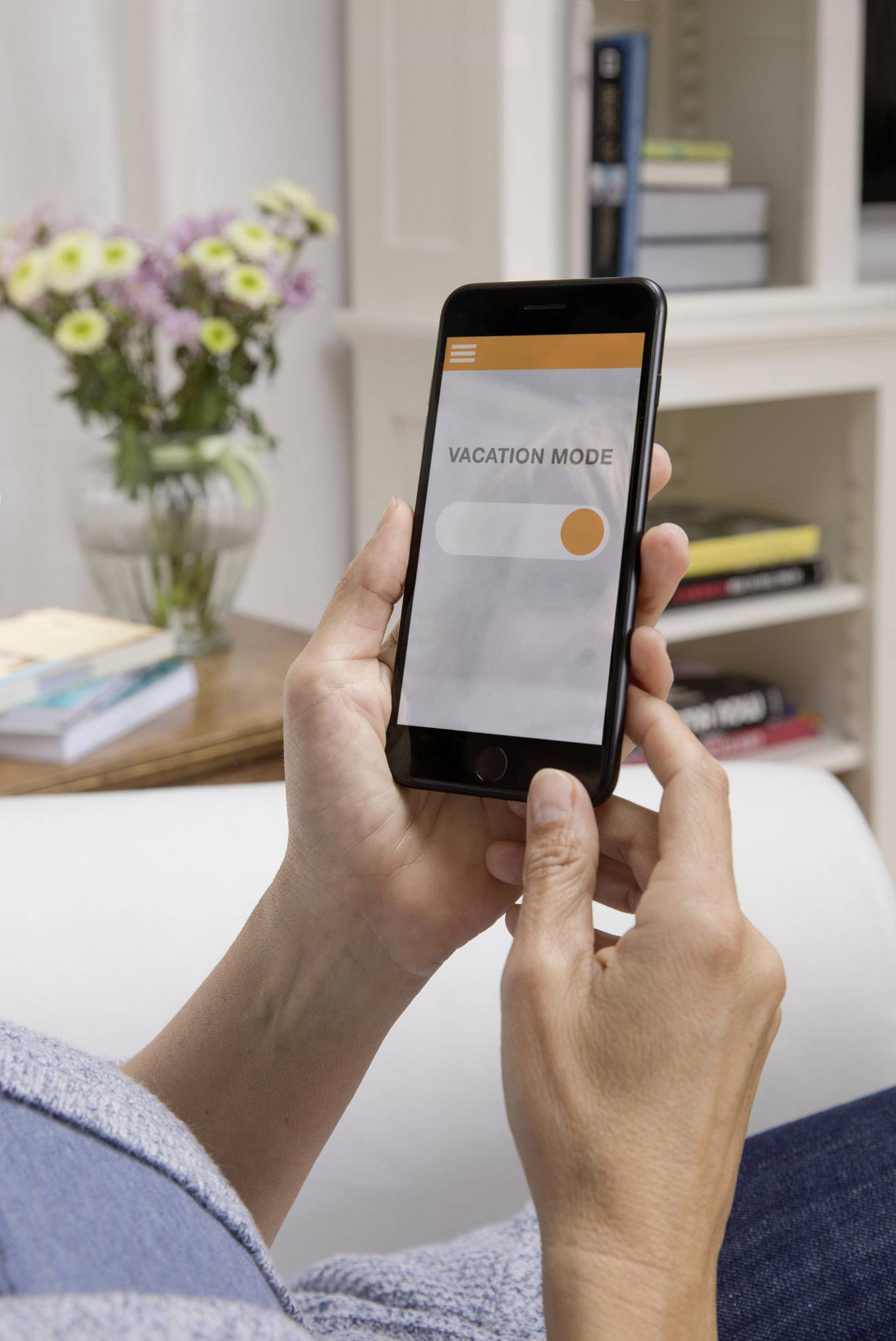 Une personne tient un smartphone affichant 'Mode Vacances'. À l'arrière-plan, des livres et un vase de fleurs sont posés sur une table.