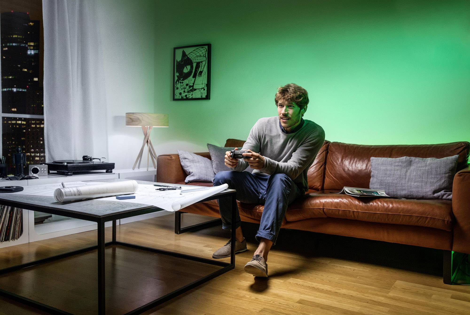 Un homme est assis sur un canapé dans un salon confortable, joue à des jeux vidéo et tient une manette. Des lumières urbaines sont visibles en arrière-plan.
