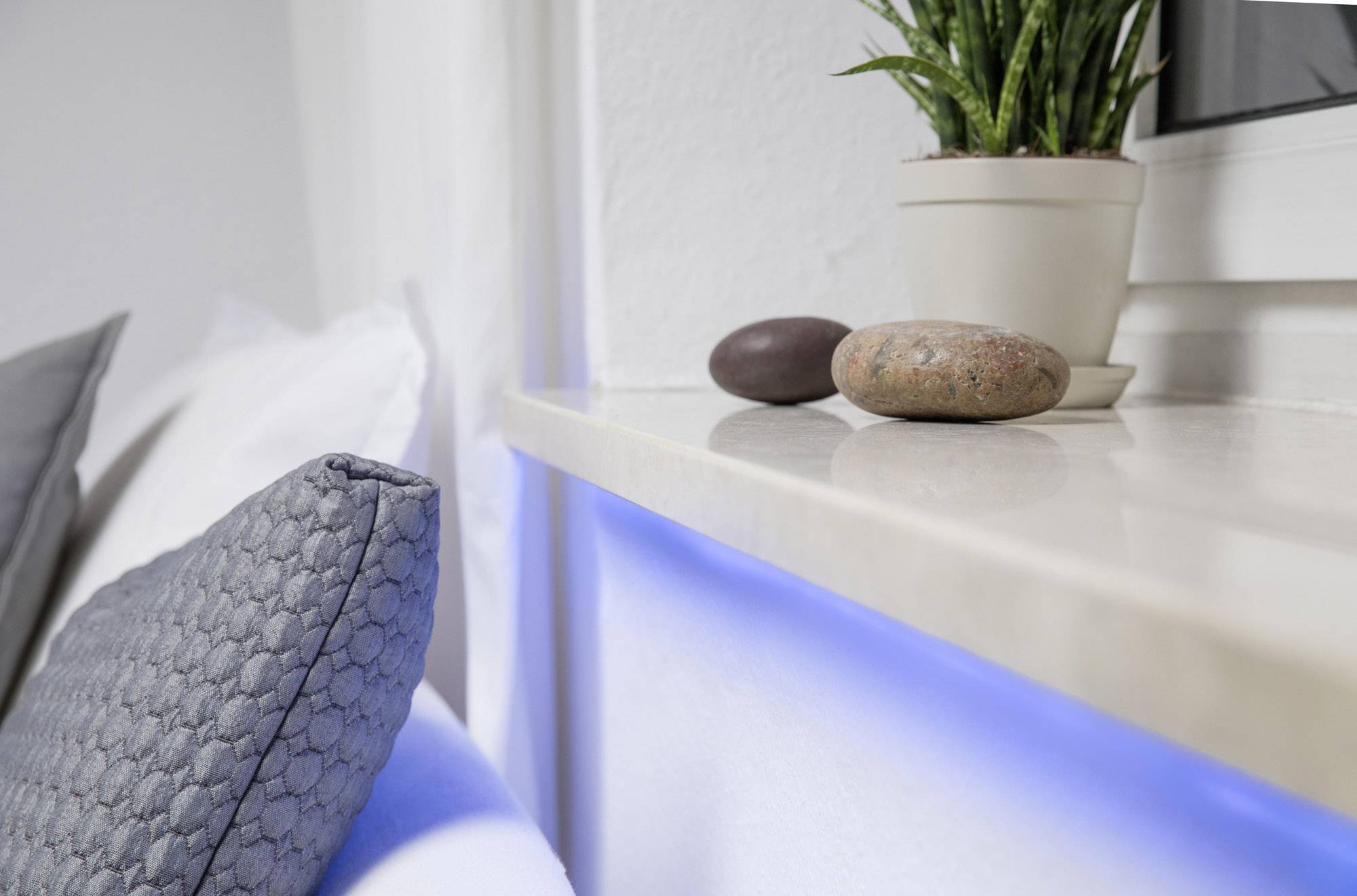 Rebord de fenêtre avec une plante en pot et deux pierres décoratives. À côté, un coussin, avec une faible lumière bleue en dessous.