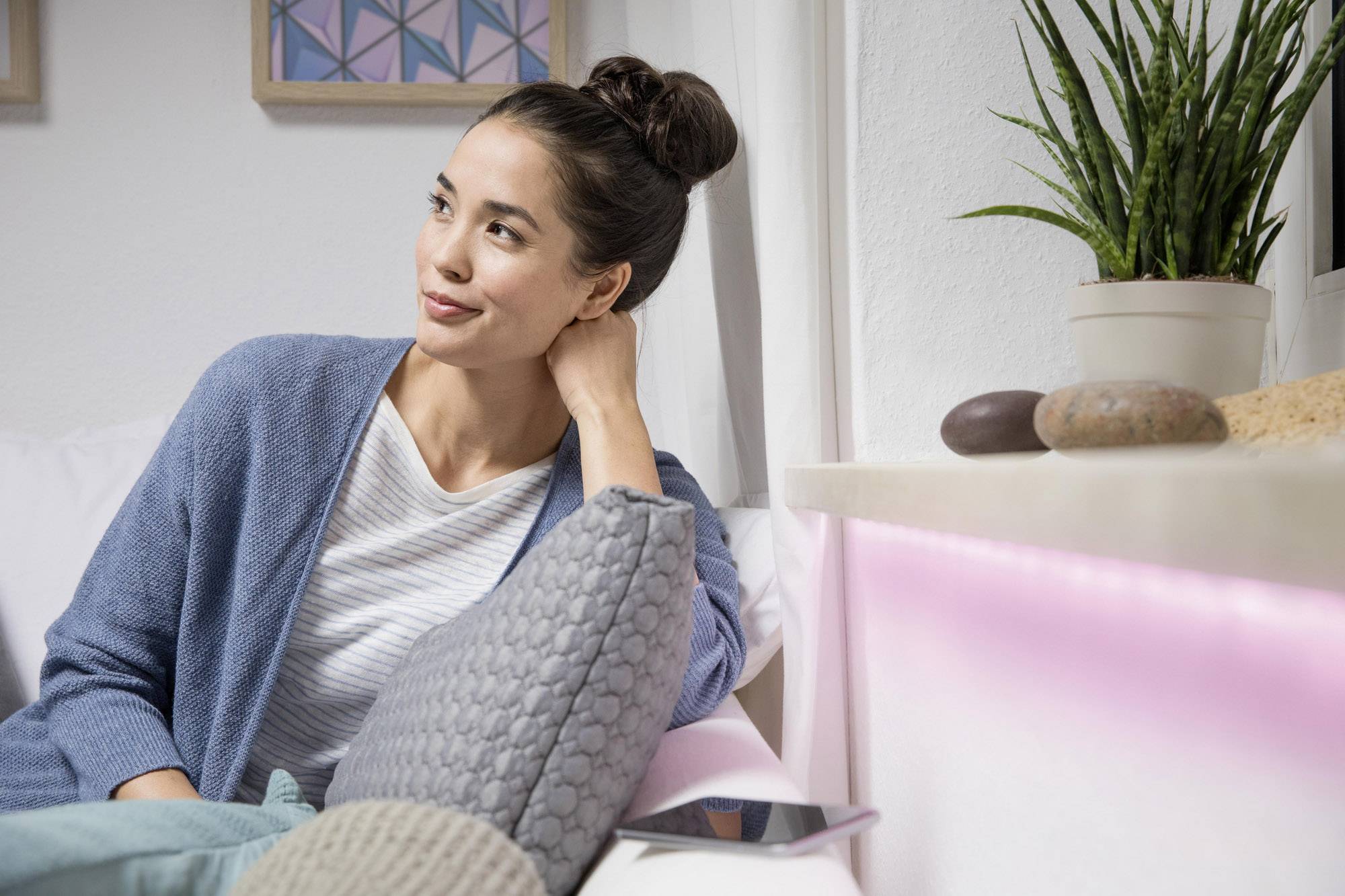 Une femme est assise tranquillement sur un canapé, souriant en regardant sur le côté. À côté d'elle, des plantes et des décorations sont posées sur une étagère.