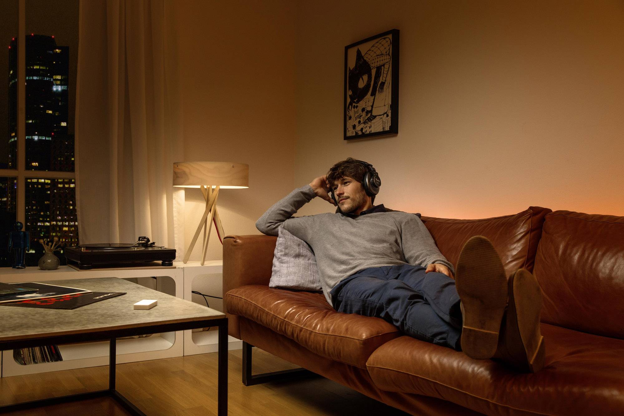 Un homme est allongé de manière détendue avec des écouteurs sur un canapé en cuir brun dans un salon doucement éclairé avec vue sur la ville.