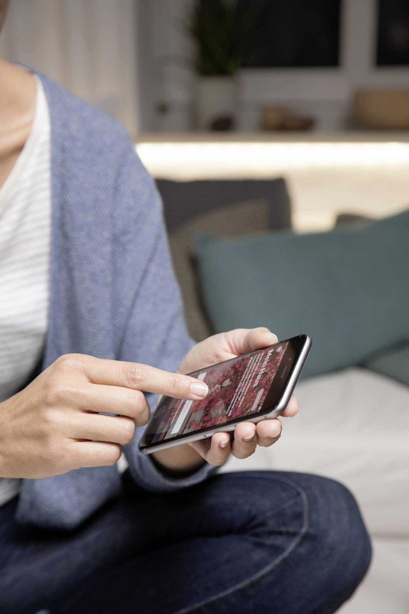 Une personne fait défiler un smartphone dans un espace confortable, probablement un salon avec des coussins en arrière-plan.