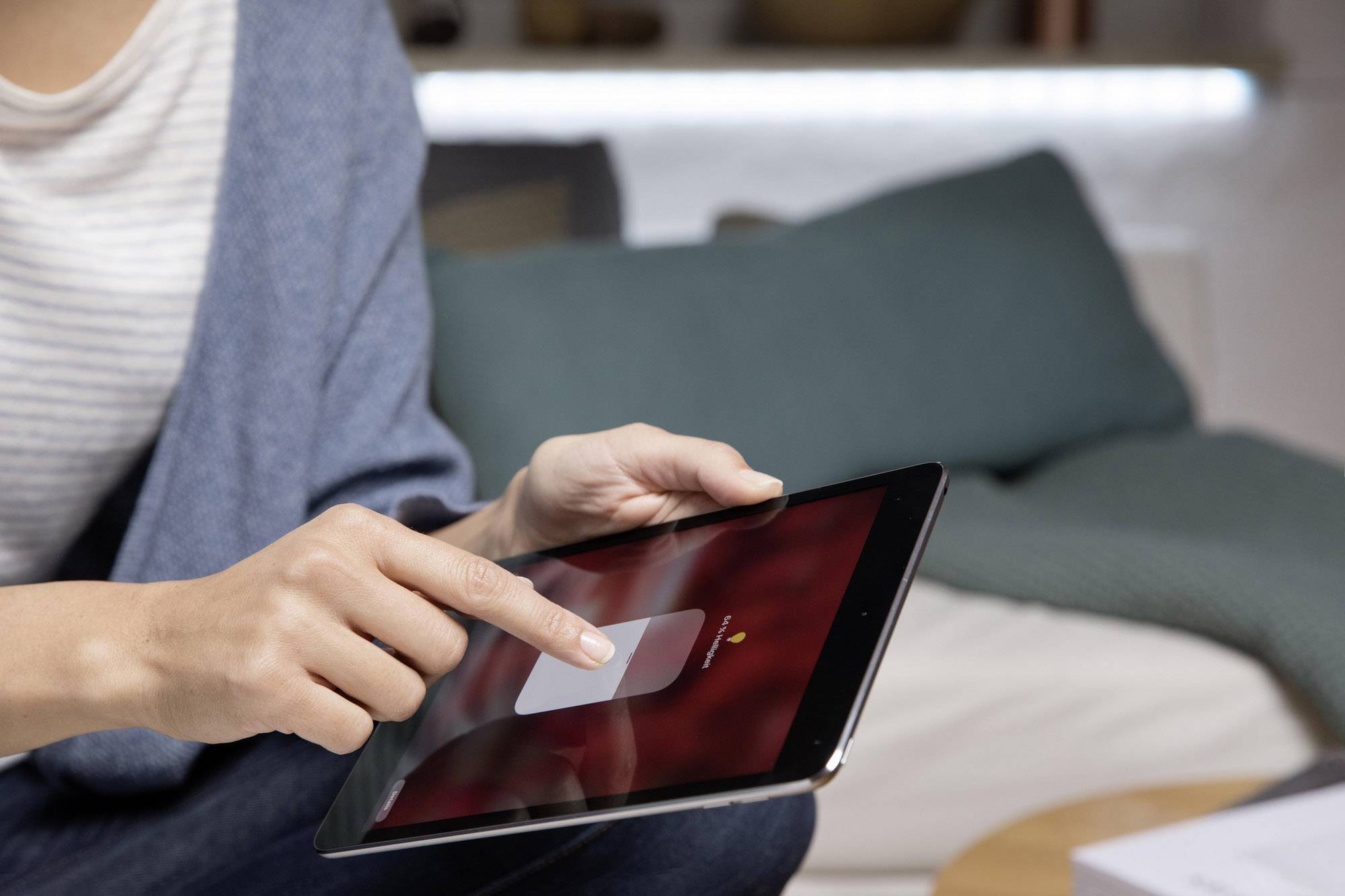 Une personne utilise une tablette avec son doigt depuis un siège de canapé. L'environnement semble confortable, avec des coussins en arrière-plan.