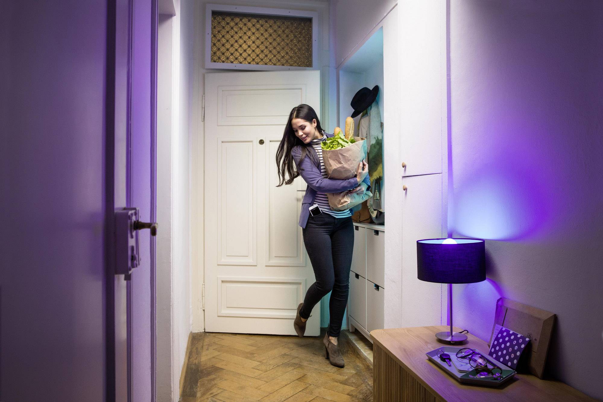 Une personne entre dans un couloir avec un sac de courses rempli de nourriture, sourit et porte un blazer, un chapeau et une écharpe en arrière-plan.