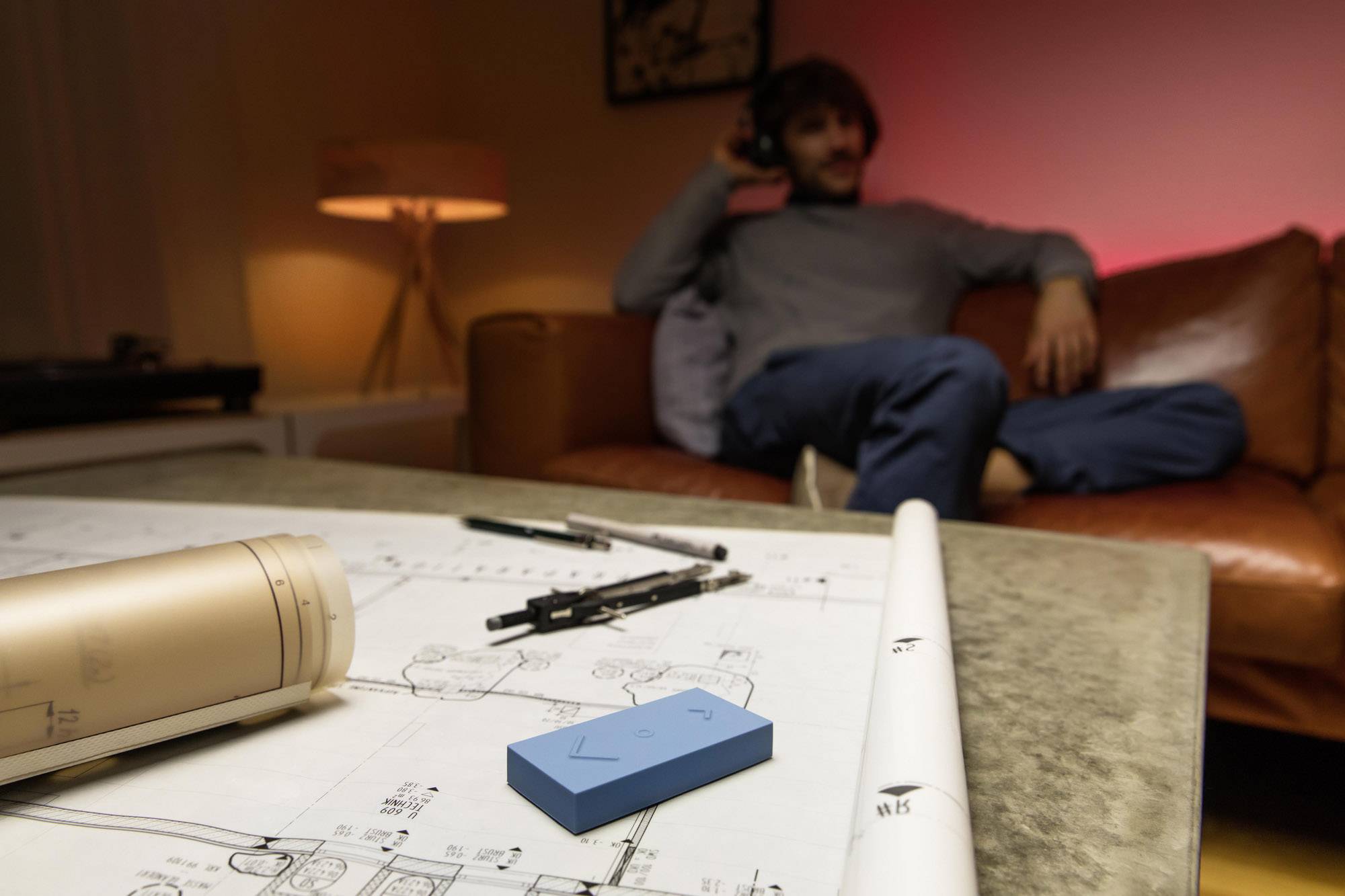 Esquisses d'architecture sur une table avec un outil de planification au premier plan ; en arrière-plan, une personne floue est assise sur un canapé.