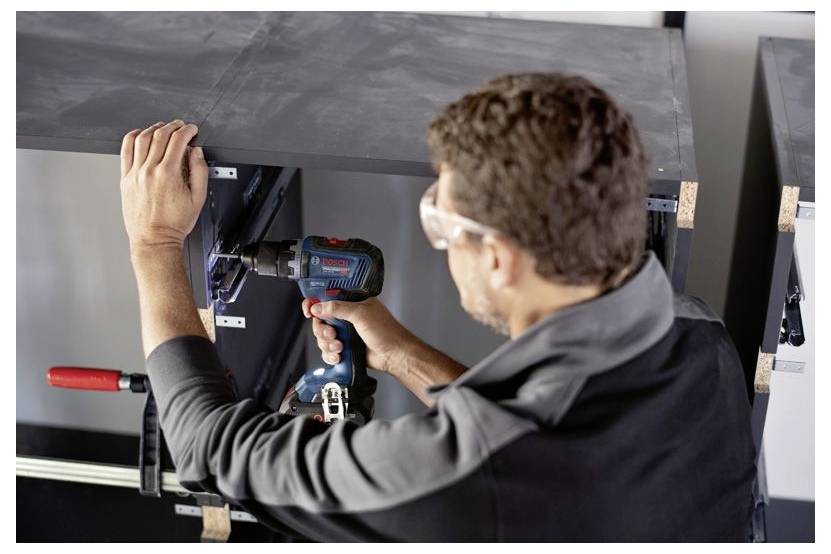 Une personne utilise une perceuse électrique pour assembler un meuble noir, portant des lunettes de sécurité. Le meuble est maintenu en place avec des serre-joints.