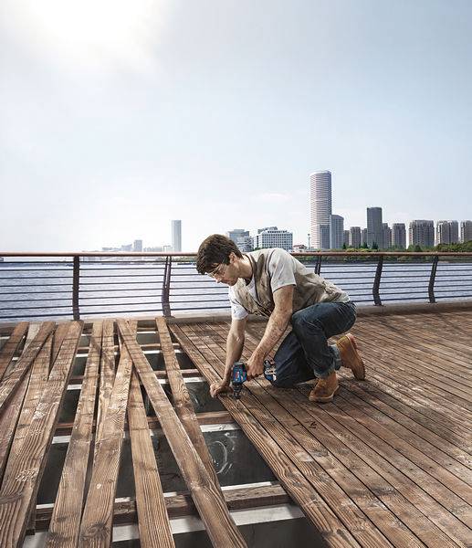 Un homme rénove une terrasse en bois près de l'eau. Des immeubles de grande hauteur d'une ville sont visibles en arrière-plan.