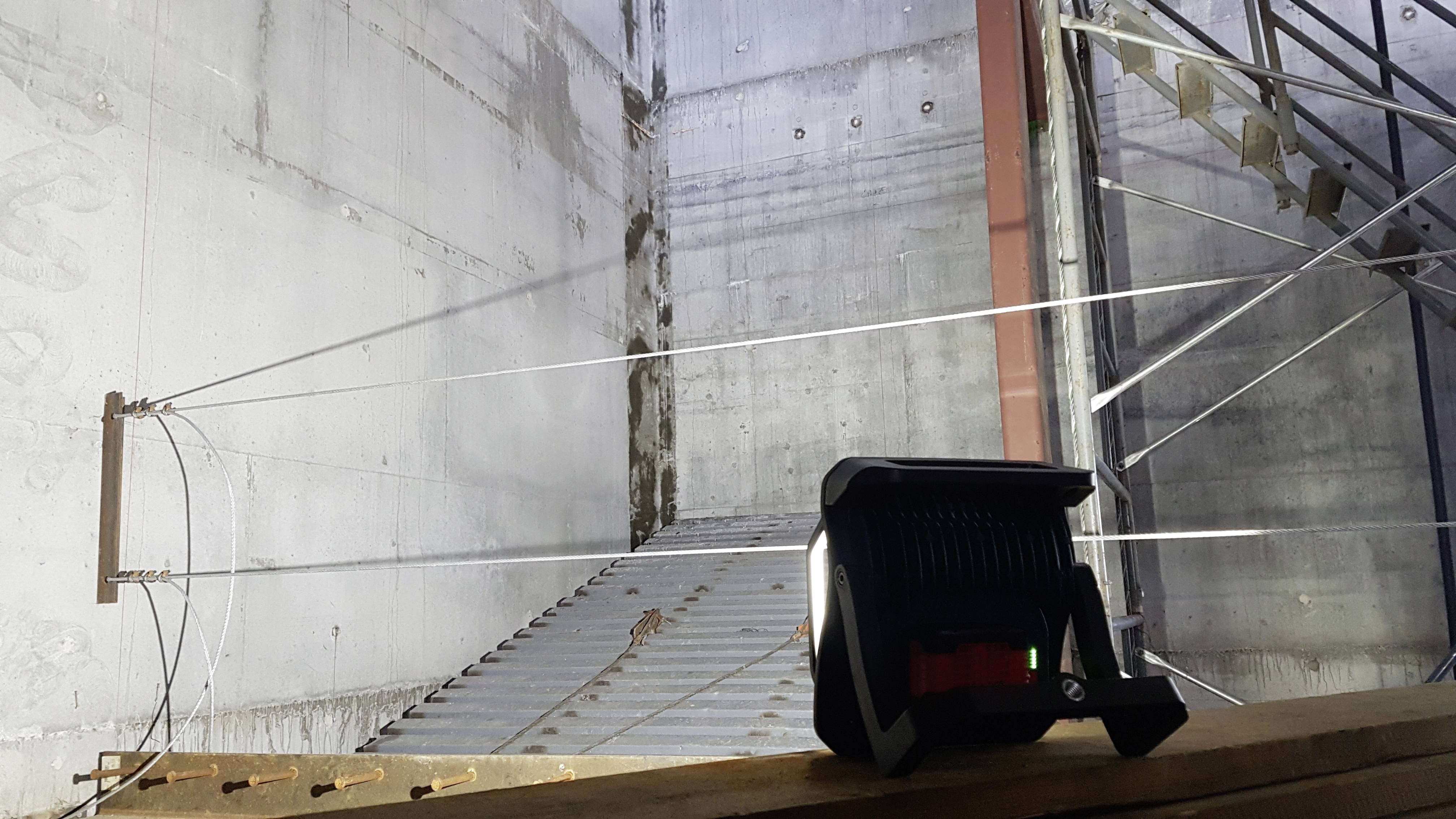 Un chantier à l'intérieur d'un bâtiment. Un projecteur de chantier éclaire un mur. Des échafaudages métalliques et des escaliers sont visibles.