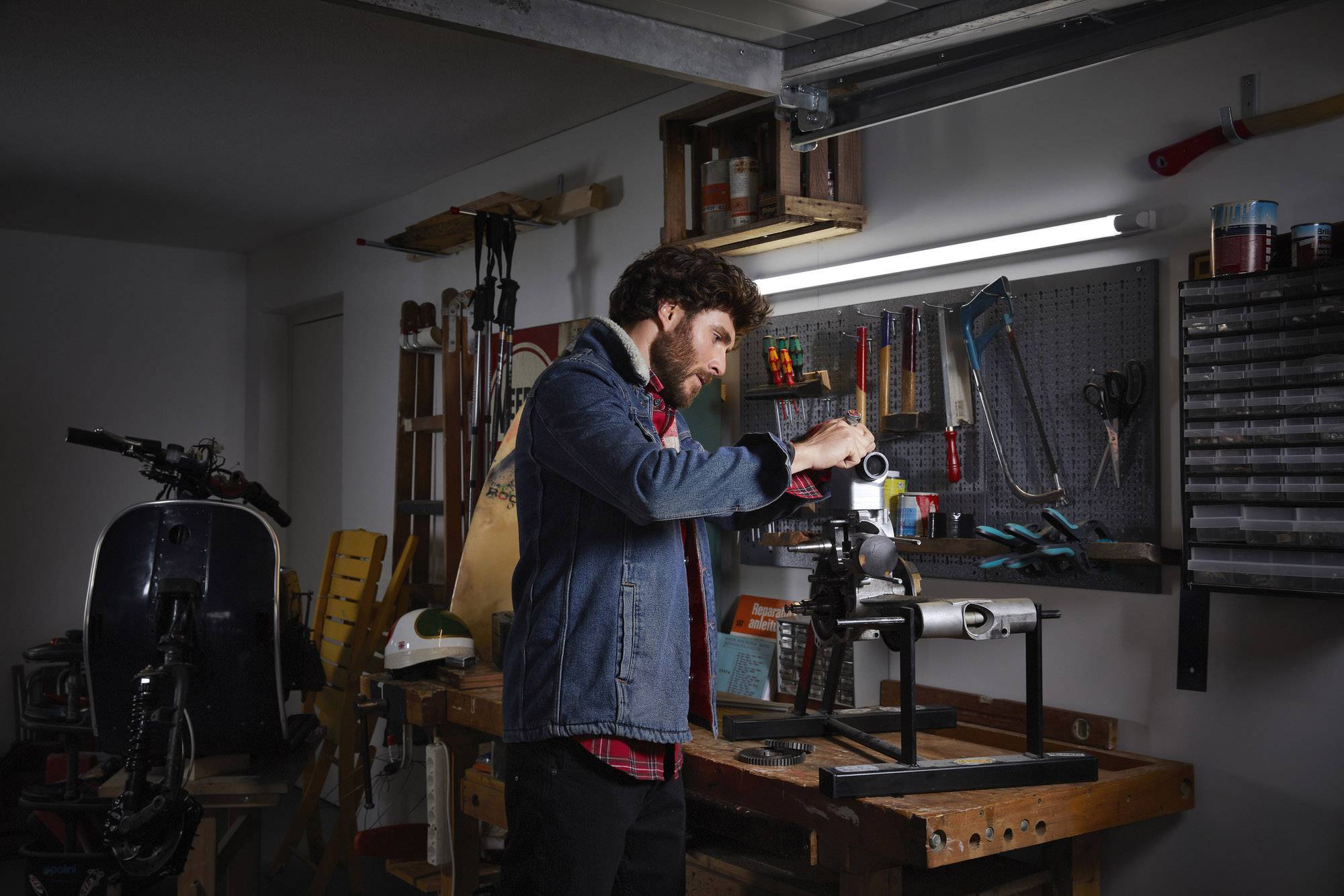 Un homme travaille dans un atelier, entouré d'outils et de machines. Il porte une veste en jean et se concentre sur un projet.