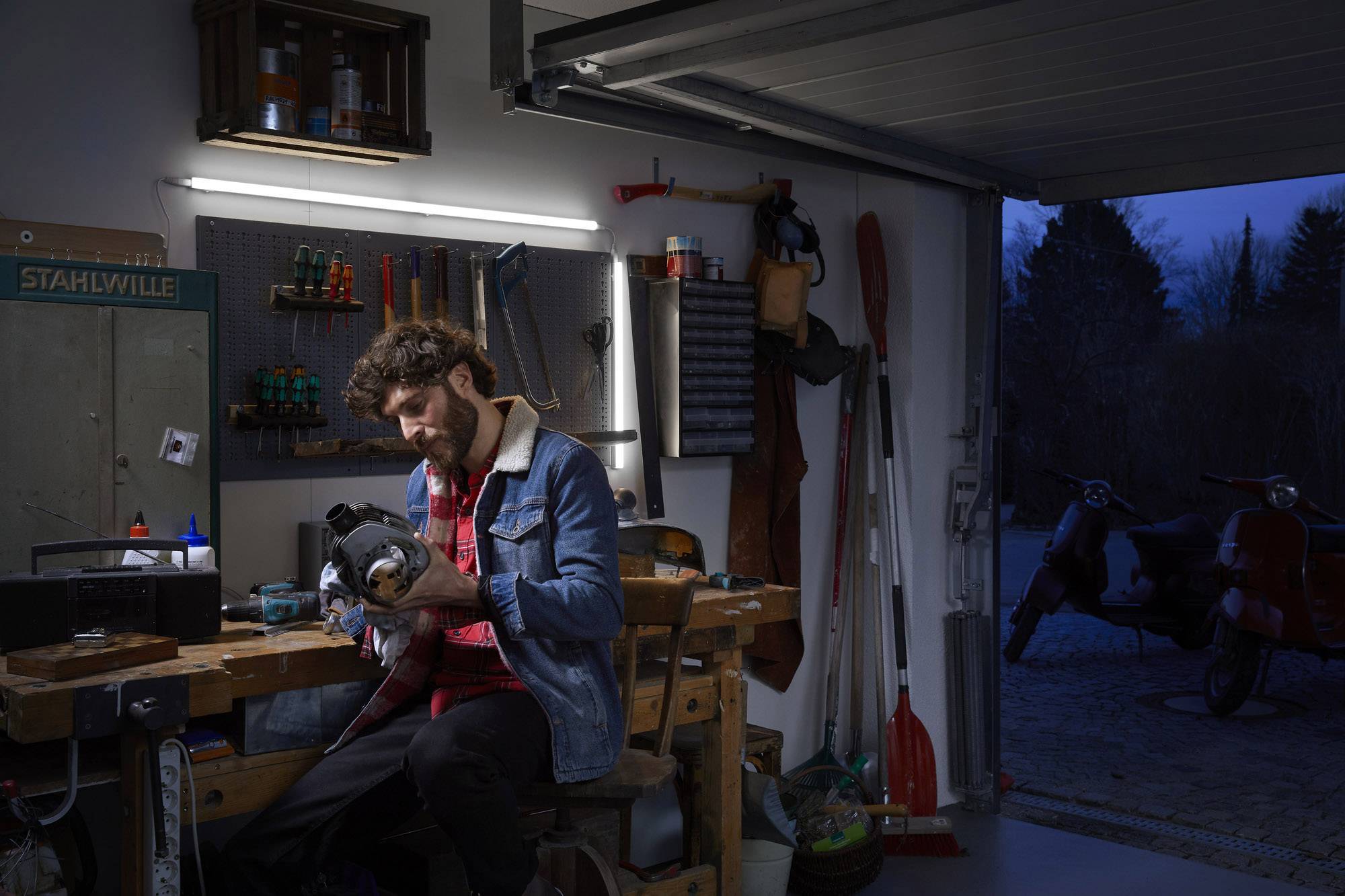 Un homme est assis à un établi dans un garage, absorbé par son travail avec un outil, alors qu'il fait sombre dehors.