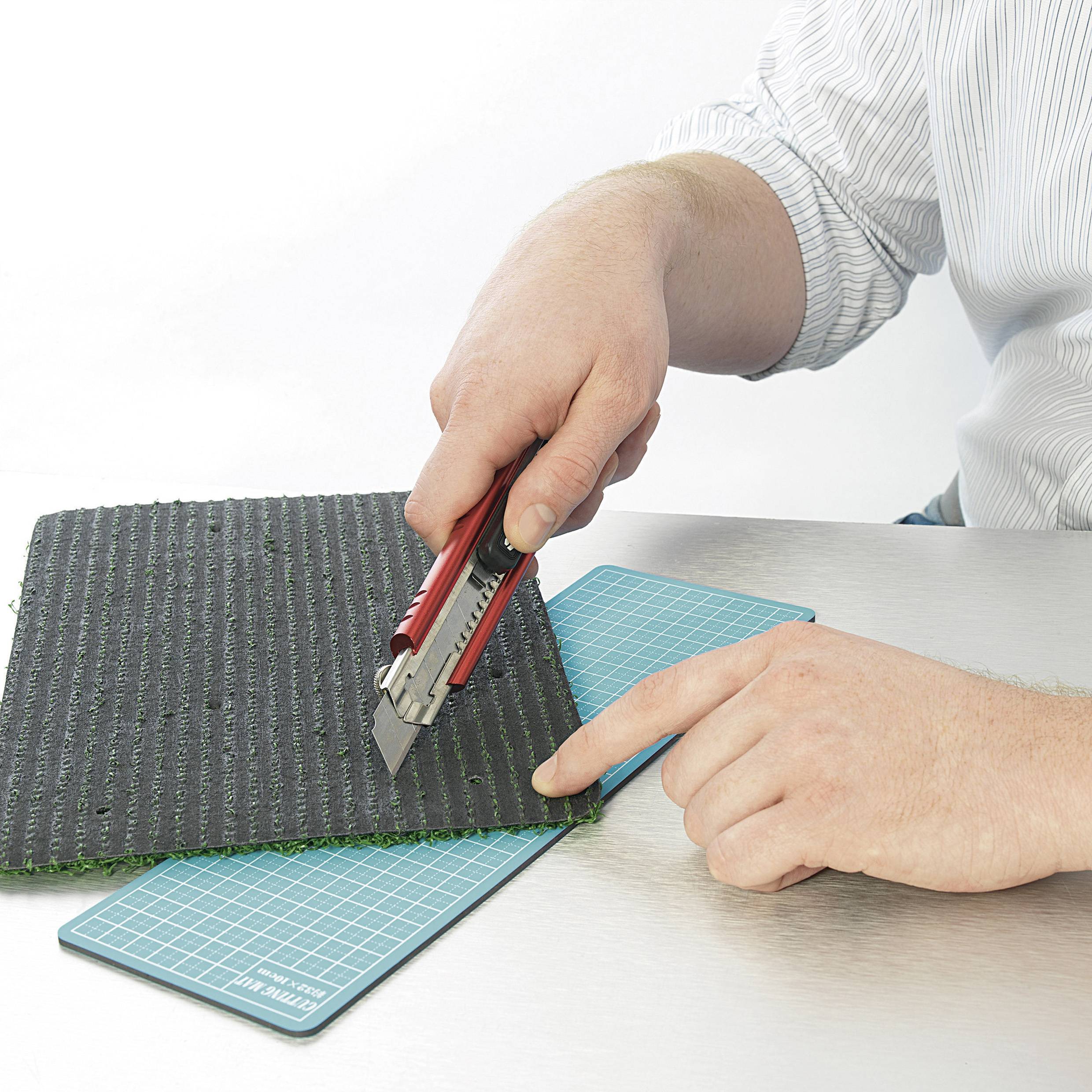 Une personne coupe un morceau de gazon artificiel avec un cutter rouge sur un tapis de coupe.