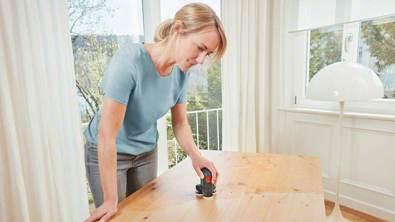 Une femme utilise un petit appareil portatif pour travailler la surface d'une planche en bois dans une pièce lumineuse et moderne.