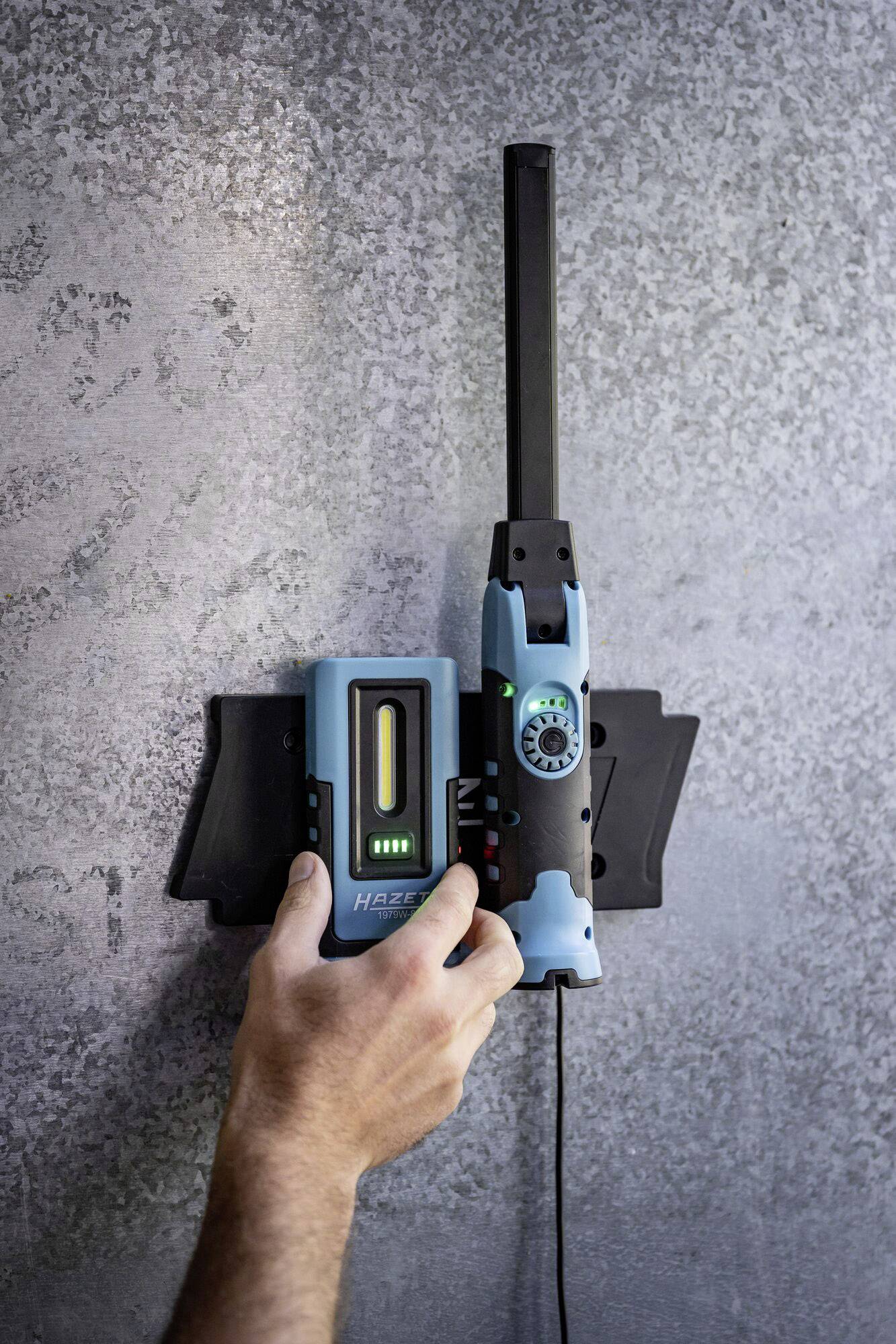 Une main utilise un appareil bleu et noir pour détecter des objets à travers un mur. L'appareil affiche un écran lumineux.