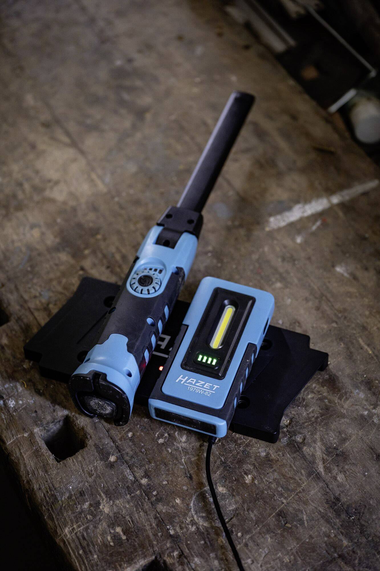Une lampe de travail rechargeable bleu-noir est posée sur sa station de charge sur un établi en bois. Elle est équipée de LED indiquant son niveau de charge.