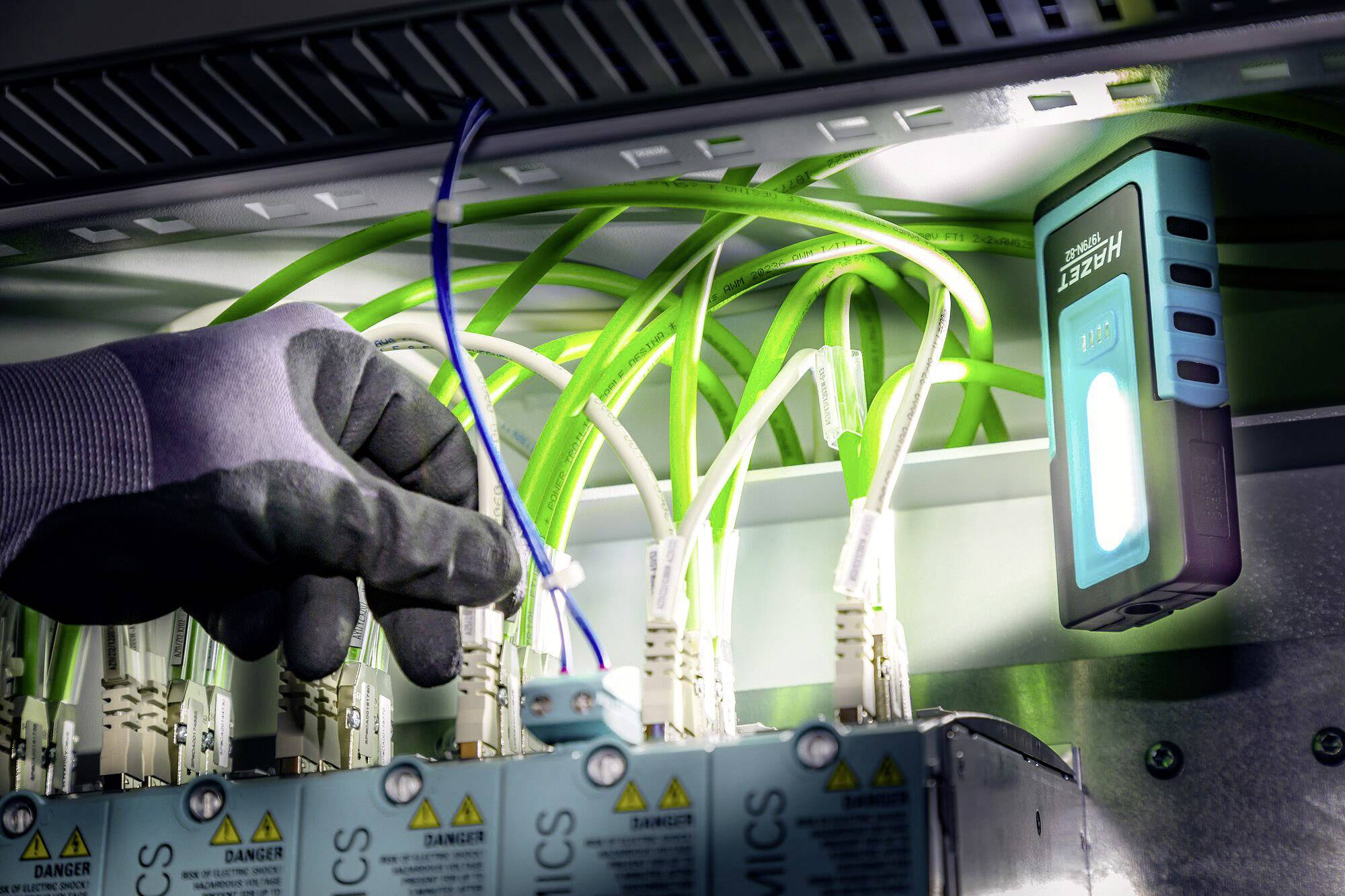 Une main gantée travaille sur des câbles réseau ou à fibre optique dans une salle de serveurs, accompagnée d'un appareil de test lumineux.