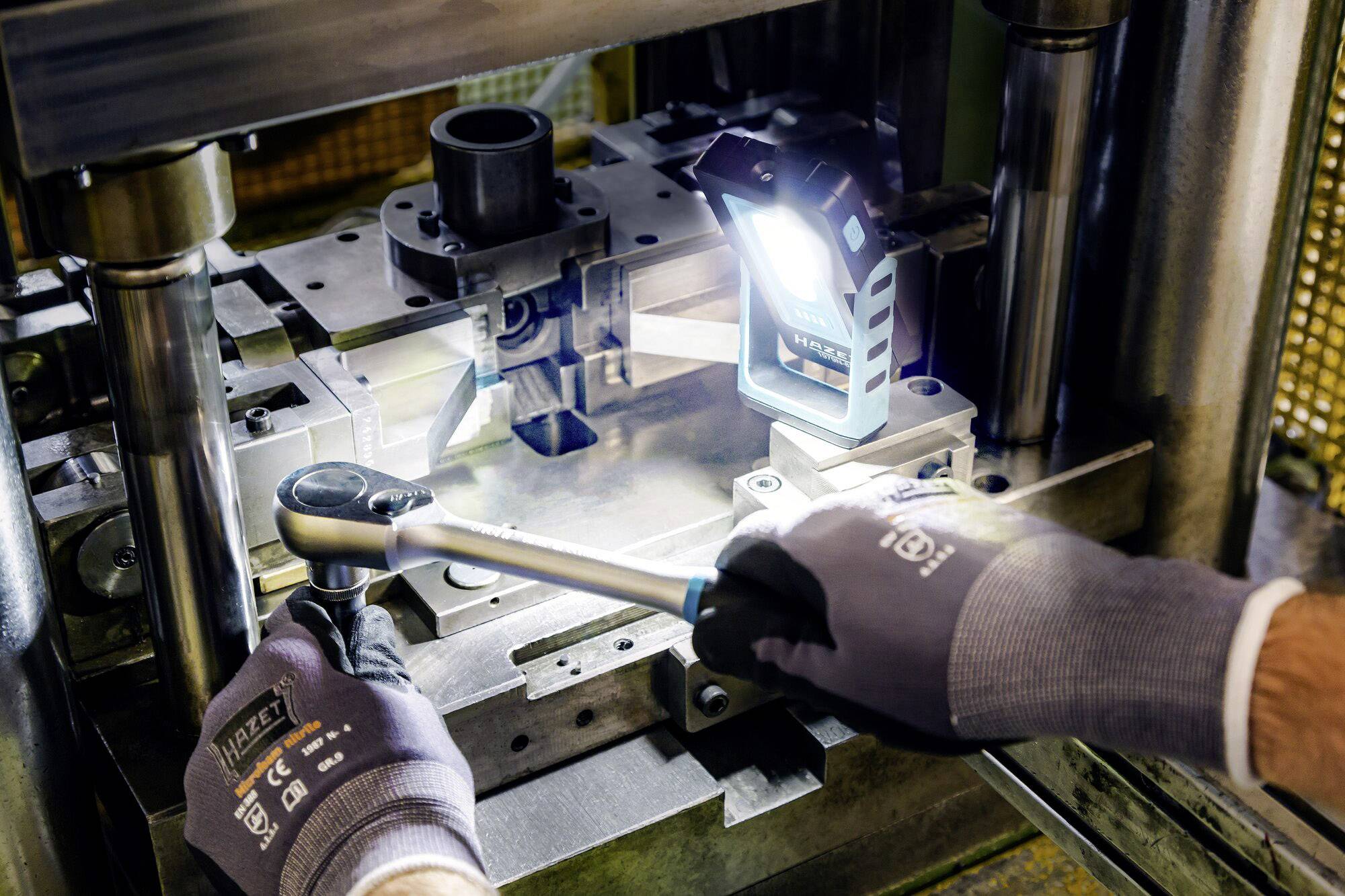 Une personne portant des gants de travail ajuste une machine métallique à l'aide d'une clé, tandis qu'un projecteur de chantier éclaire la zone.