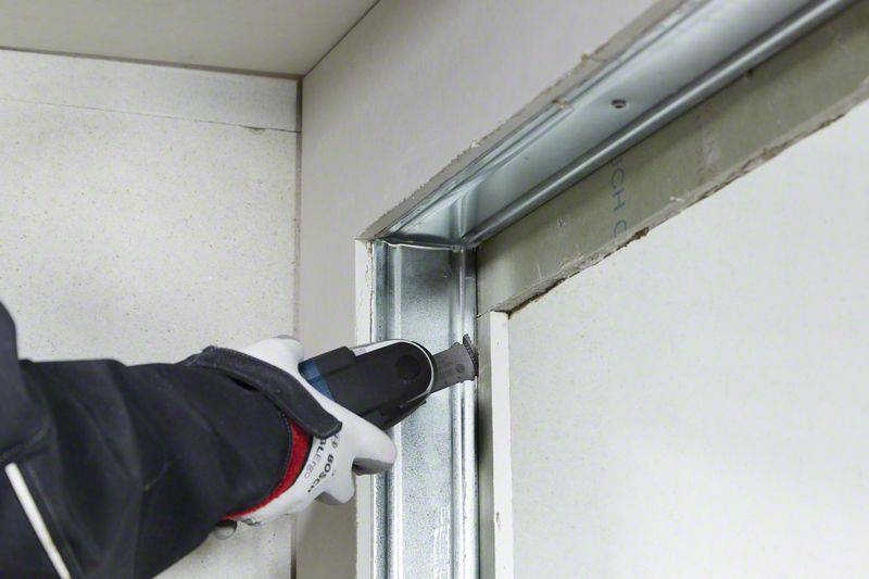 Une main coupe une cloison en plaque de plâtre avec un cutter. Le cadre en profilé métallique est visible. Un gant de protection est porté à la main.