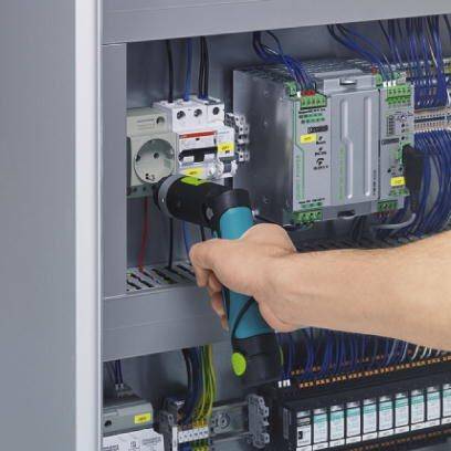 Un homme examine un tableau de commande à l'aide d'un appareil électronique. Plusieurs fils et composants sont visibles.